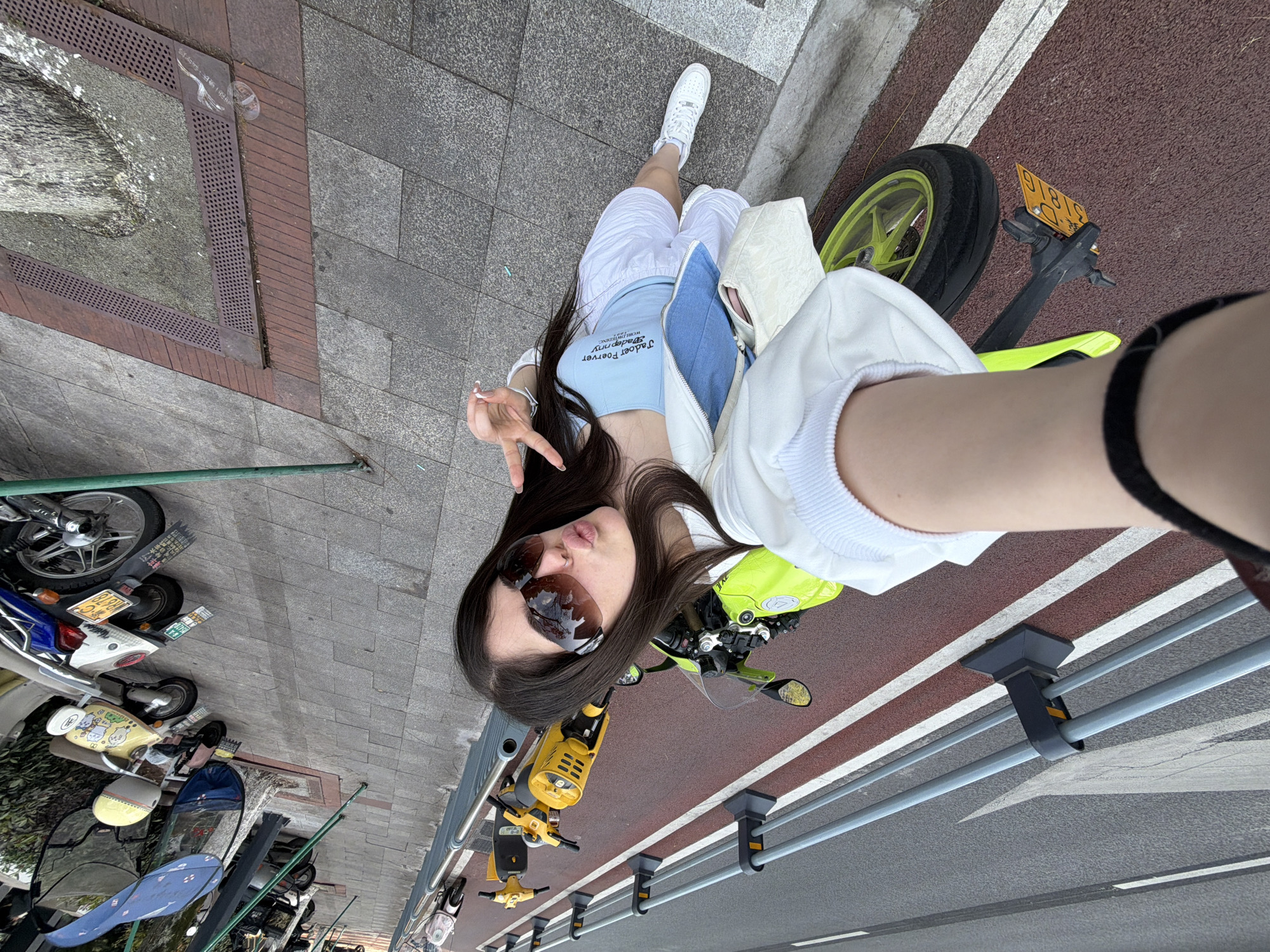 The image is a high-angle selfie taken by a young woman in an urban setting, rotated approximately 90 degrees clockwise.
The main subject is a young woman with long, dark hair, wearing large sunglasses, a light blue crop top, a white zip-up jacket, and white shorts. She has a nose piercing and is making a peace sign with her left hand while pouting her lips. Her white sneakers are visible. The text "Jadoo Forever" and "WORLD FASHION" is visible on her top.
She is positioned next to a bright lime green sport motorcycle. A yellow license plate on the motorcycle reads "D-31816". A light-colored shoulder bag is resting on the motorcycle's seat. In the background, various scooters and bicycles are parked along the sidewalk. One yellow scooter features cartoon characters, and a white scooter has a license plate "86G70" with the text "平安出行" (Safe travel) on its holder.
The setting is a city sidewalk made of grey paving stones, with sections of red brick and a metal drain grate. Faint Chinese characters on the ground near the drain appear to say "注意安全 保持距离" (Pay attention to safety, keep distance). Adjacent to the sidewalk is a road with a distinct red lane, likely for bicycles or scooters, marked with white lines, and a grey asphalt section. A metal railing separates the sidewalk from the red lane.
The activity shows the woman posing for a playful selfie. It is daytime, and the lighting suggests clear weather. The location context is Chongqing High-tech Zone, China.