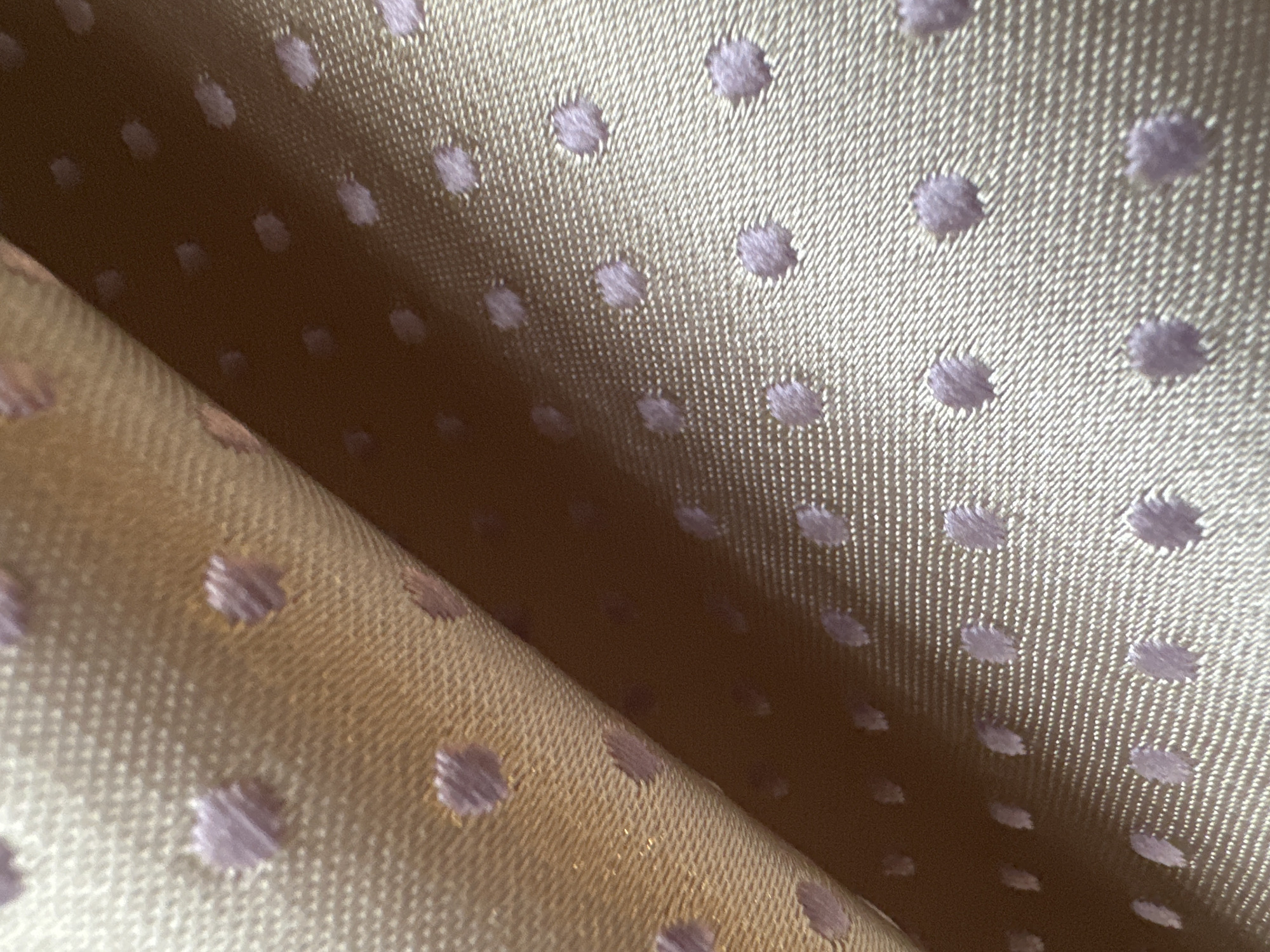 A close-up shot shows a folded piece of light beige fabric with lavender polka dots. The fabric has a visible woven texture. The lighting creates shadows and highlights that define the folds.