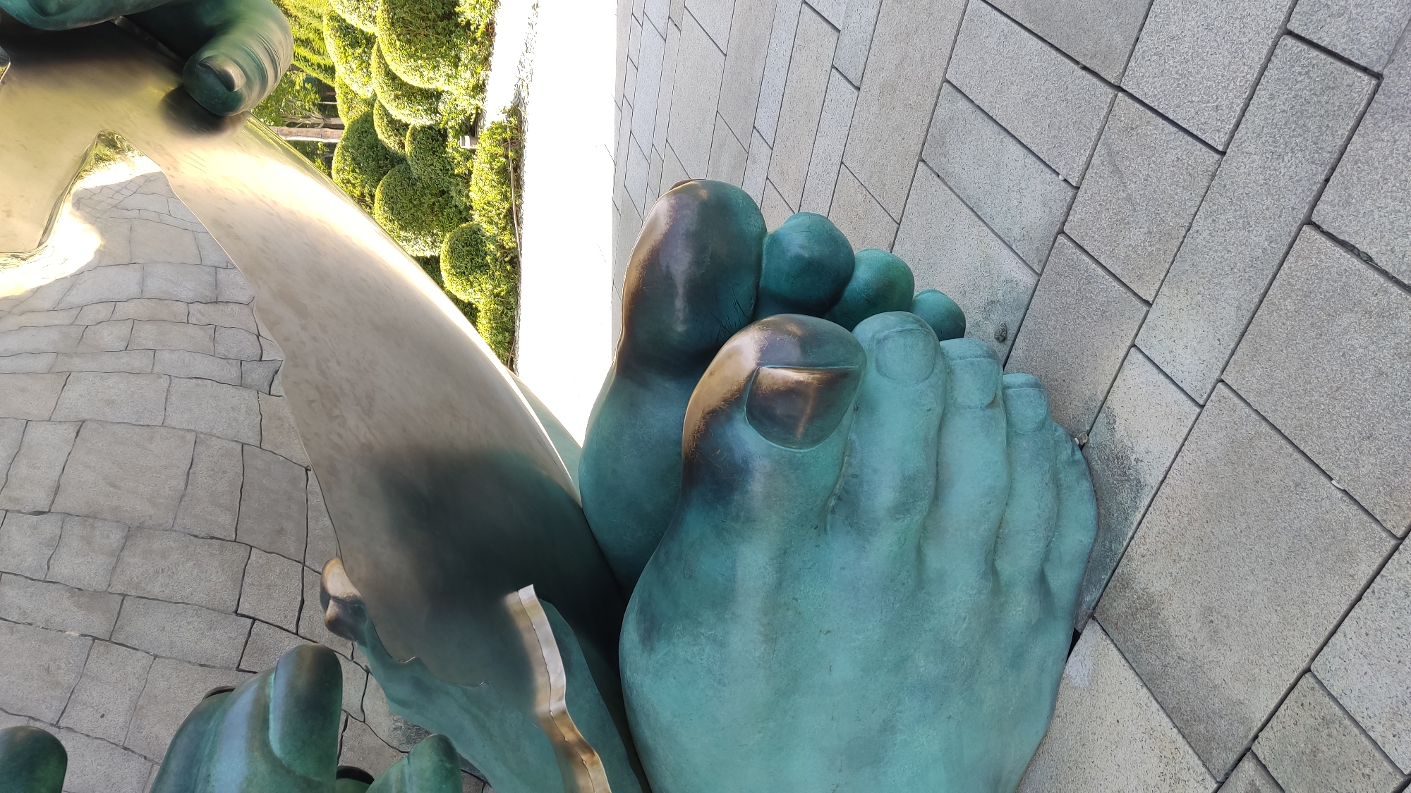 The image shows a close-up of the bronze feet of a monumental statue, likely the Monument to the Conquerors of the Kuban, located in Krasnodar, Russia. The statue's feet are weathered, with a green patina, and are positioned on a paved area composed of rectangular grey stone tiles. In the background, there are manicured green bushes and a stone wall. The lighting suggests it is daytime, with sunlight illuminating parts of the statue and the surroundings.