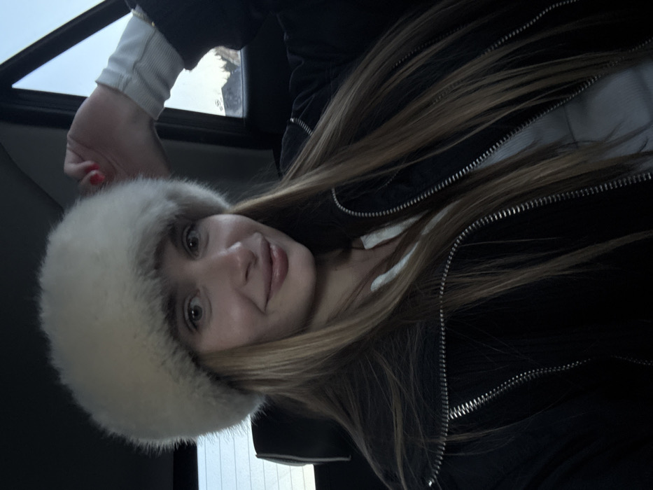 A female individual is seated in the rear passenger compartment of a vehicle. She wears a large, light-colored fur hat, a black jacket with silver zippers, and a white ribbed long-sleeved top. Her long, straight, light brown hair extends below her shoulders. She looks directly forward, exhibiting a slight smile. Her right hand, displaying red nail polish, is raised with fingers slightly curled, positioned near the vehicle's window frame. The headrest of the seat behind her is visible. Through the vehicle's side window, an outdoor scene is visible, including a light blue sky and dark shapes resembling distant trees or mountainous terrain, consistent with a winter or cold-weather environment. The image is described as being taken in Razvršje, Montenegro.