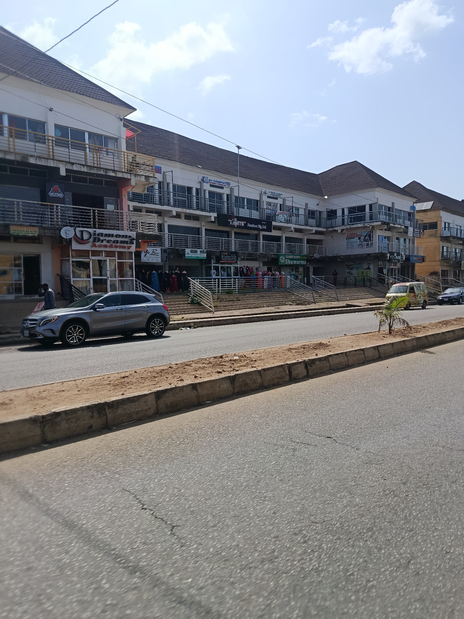 The image depicts a commercial street scene in Jos, Nigeria. A modern, multi-story building complex with a white facade and dark brown tiled roofs dominates the right side of the frame. The building features several shops on the ground floor and balconies with metal railings on the upper floors, suggesting retail and office spaces. Signs for businesses such as "Shawarma," "Diamond Dreams Academy," and "Luxury World" are visible.
In the foreground, a grey Mercedes-Benz SUV is parked on the street, facing left. A man in a grey shirt and black trousers is standing near the entrance of one of the shops. The street is paved with asphalt and separated from the sidewalk by a concrete and gravel curb. A small palm tree is planted on the curb.
Further down the street, a yellow minivan is driving away from the camera, and a dark-colored car is following it. The sky is a light blue with scattered white clouds. The overall impression is of a busy commercial area on a bright, sunny day.