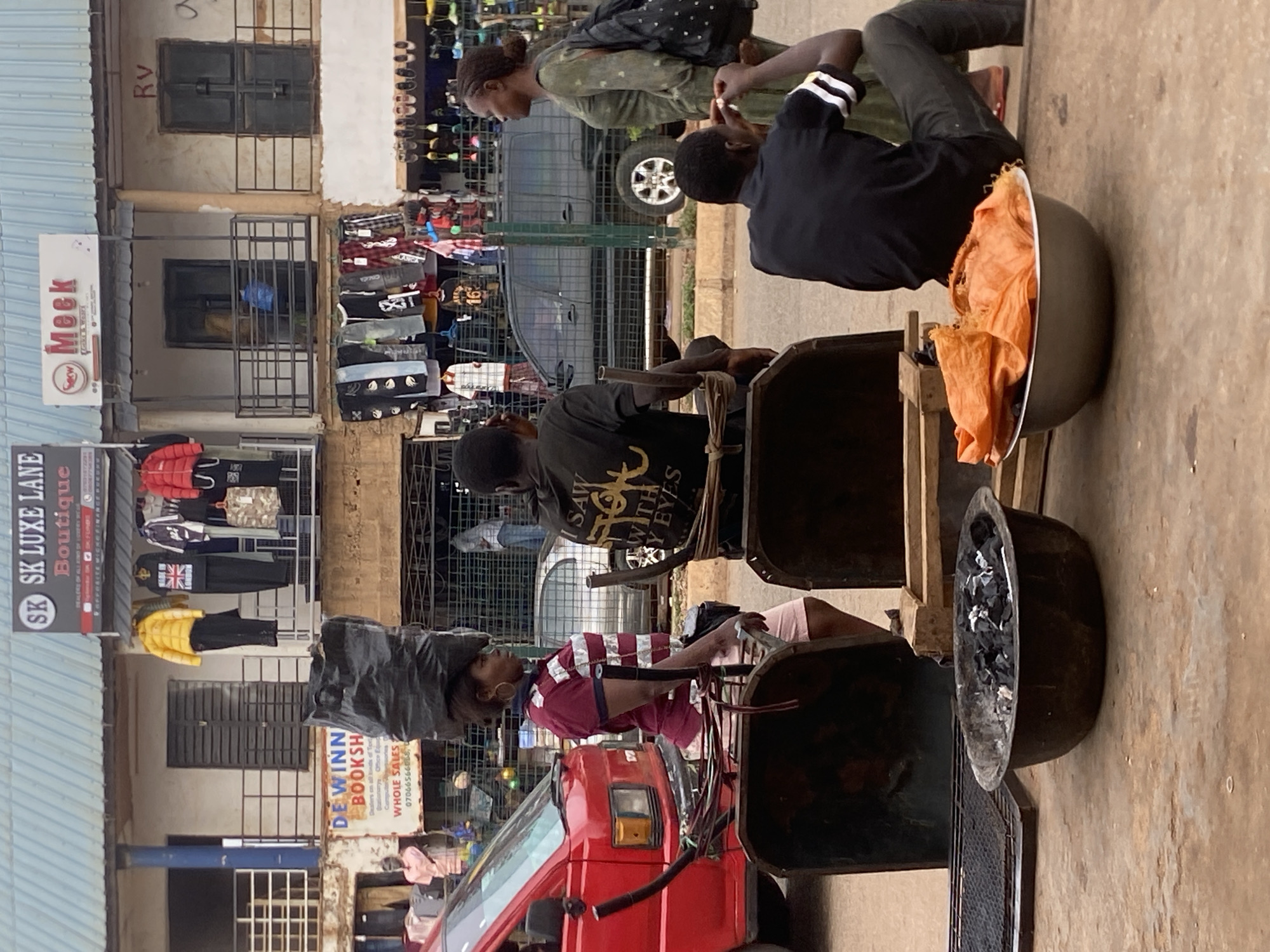 The image depicts a busy street scene likely in Bukuru, Nigeria, characterized by market stalls and street vendors. A red three-wheeled vehicle or tuk-tuk with black cables trailing is positioned in the foreground. A woman carries a large load on her head, secured by a head pad. Several people are gathered around a wooden cart containing multiple bowls or containers. One bowl holds orange textiles, while another appears to contain charcoal. Two young men are involved in a transaction, one sitting on a high stool. A clothing stall displaying various items is visible in the background, along with multiple store signs: "SK LUXE LANE Boutique," "DEWINN BOOKSH" (bookshop), and "Meck." The architecture indicates a commercial area with buildings two to three stories high. The street surface is light-colored and shows signs of use. The lighting suggests it is daytime.
