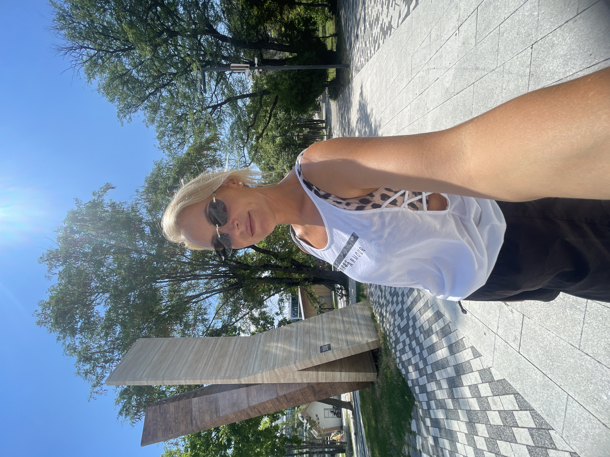A woman with blonde hair and sunglasses is taking a selfie outdoors on a sunny day. She is wearing a white tank top with leopard print accents and black shorts. Behind her, there is a modern sculpture made of large, angular wooden and stone panels. The sculpture is situated in a park-like area with trees and a paved walkway. The walkway has a distinctive black and white checkered pattern. In the background, there are more trees, buildings, and streetlights. The location is the park next to the "Memory of the Siege of Novorossiysk" memorial, which is located on Tsezarsky Hill in Novorossiysk, Russia. The memorial itself is not fully visible in the frame.