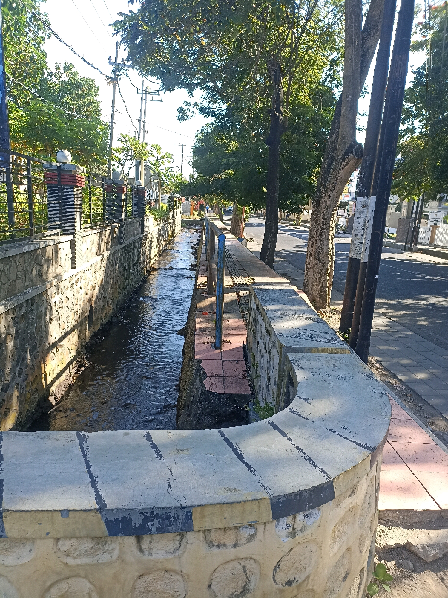 The image shows a canal running alongside a sidewalk and a street in Mojopanggung, Indonesia. The canal has stone walls and is filled with flowing water. Trees line the sidewalk, casting shadows on the street. Utility poles and wires are visible in the background. The scene appears to be an urban environment during daylight hours.