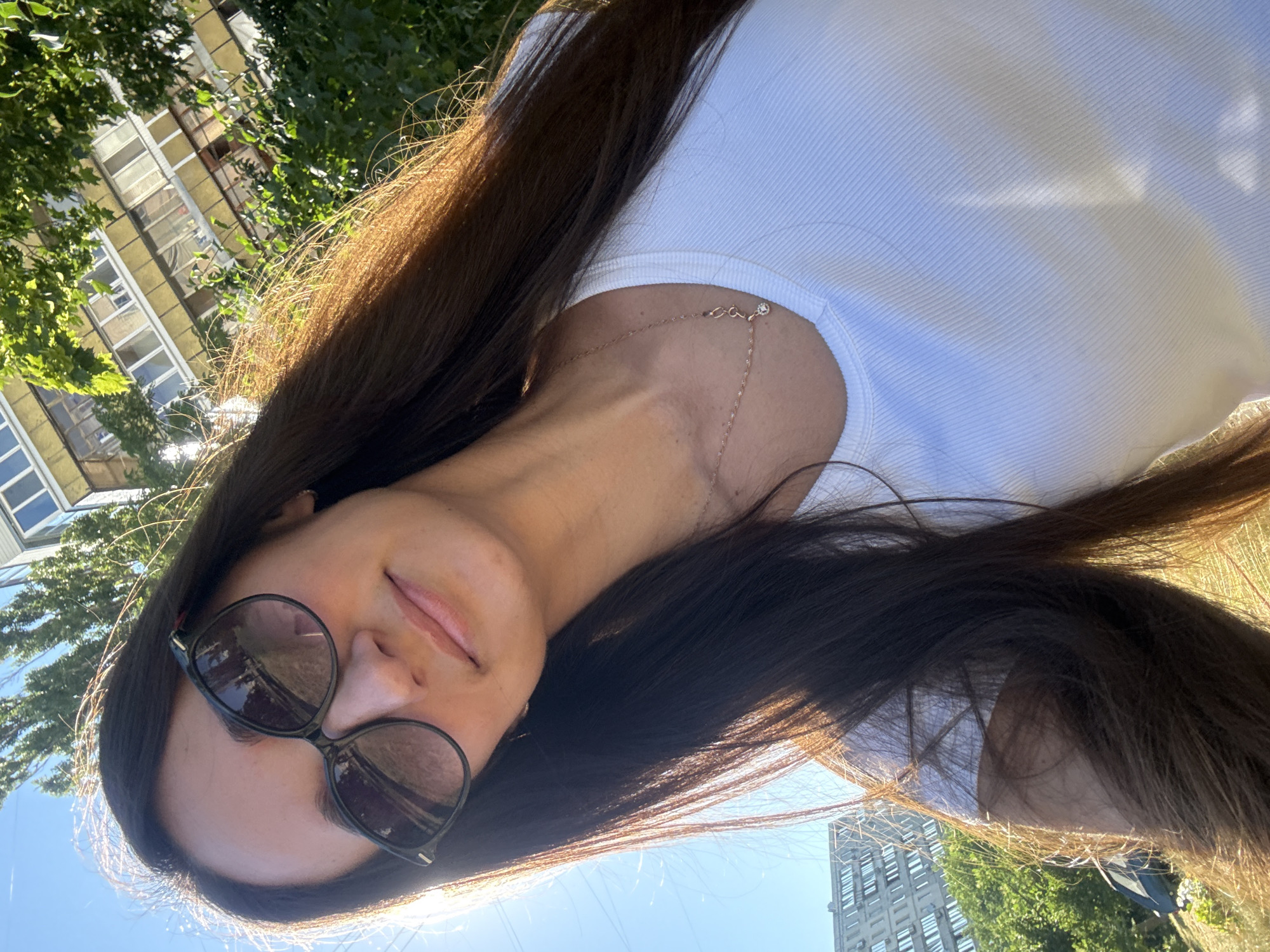 A medium shot captures a young woman with long, dark hair and sunglasses posing for a selfie outdoors on a sunny day. She is wearing a white ribbed t-shirt and a delicate gold necklace with a small pendant. Her left shoulder is slightly raised, and she is looking towards the camera with a subtle smile.
In the background, a multi-story building with yellow balconies and rows of windows is visible, partially obscured by lush green foliage. The sky is a clear blue. To the left of the frame, another building, appearing to be a residential block, is seen in the distance. The overall setting suggests an urban environment in Kyiv, Ukraine, possibly a park or residential area with trees and buildings. The sunlight creates a warm glow, highlighting the woman's hair and casting shadows. The image appears to be taken at eye level, with the woman's head tilted slightly back.