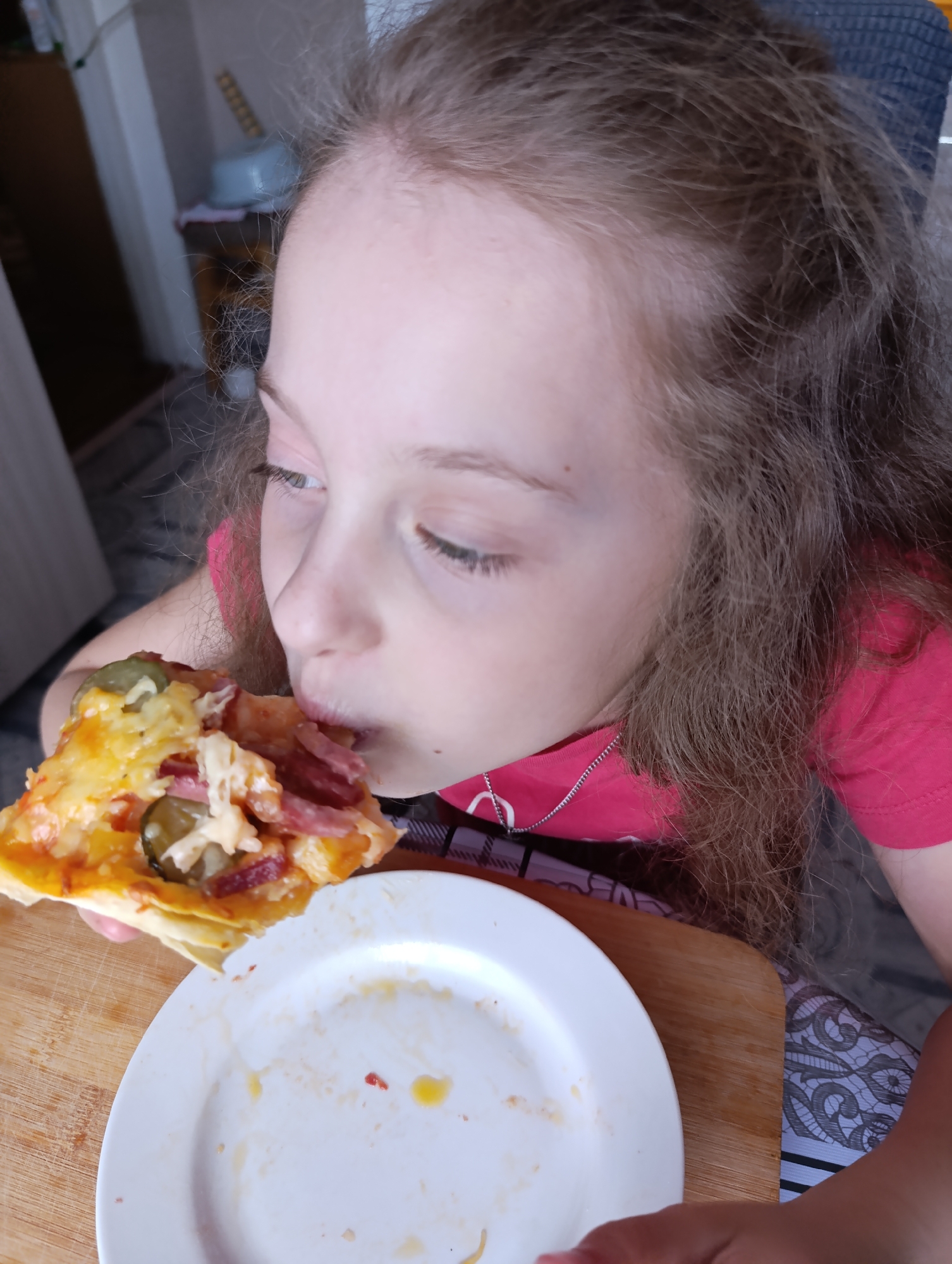 A young girl is shown in close-up, indoors, with a slice of pizza being brought to her lips. The pizza contains cheese, sausage, and green pickles. She wears a pink t-shirt and a necklace, and the remains of a previous slice is visible on a white plate. The setting appears to be a dining area with a wooden cutting board under the plate. The location is Unknown, Unknown.