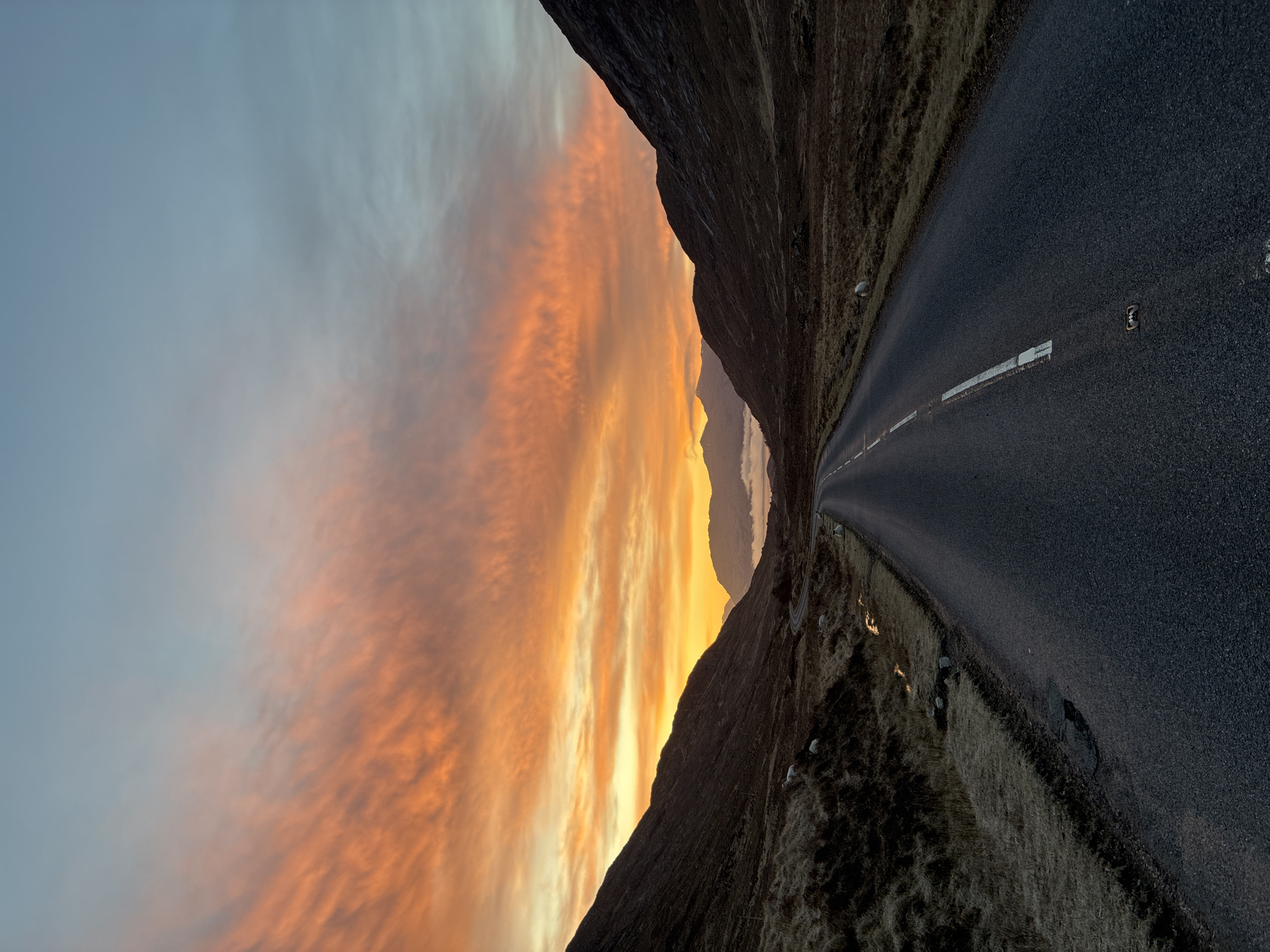 The image depicts a road running through a mountainous landscape at sunset. The road surface is dark asphalt with a painted white line in the center. On either side of the road, there are grassy slopes rising into mountains. The sky is a mix of blue and orange, with the sunset casting a warm glow on the clouds. The location is likely in the vicinity of Fort William, United Kingdom, given the terrain and lighting conditions. There are no visible people or subjects.