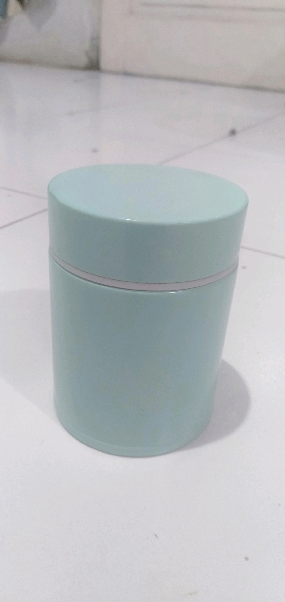 A light teal cylindrical container with a matching lid sits on a white tiled surface. A thin, light grey band encircles the container just below the lid. The background consists of a white wall with a subtle vertical line, possibly indicating a door or seam. The lighting suggests an indoor environment, with the shadow of the container cast to the right. The image appears to be taken at a slightly low angle. The scene is devoid of any people or signs of activity, suggesting a still life composition. No specific landmarks or features can definitively place this image within Cikoneng, Indonesia; it depicts a generic interior setting.