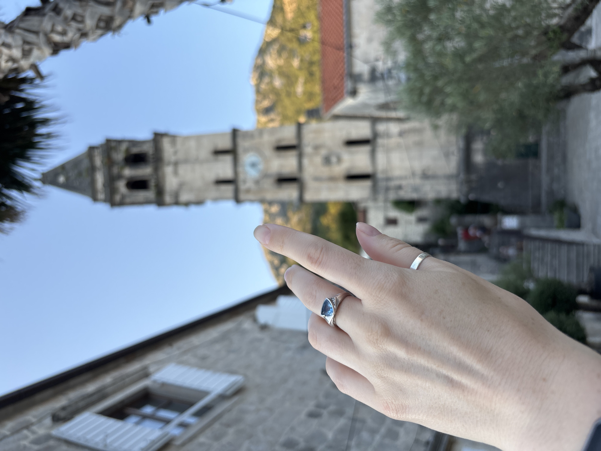 A close-up, eye-level, outdoor, medium shot captures a person's left hand with two silver rings. The hand is positioned in the foreground, obscuring the lower left quadrant of the image. The index finger is extended upwards, pointing towards a tall, stone bell tower in the background. The bell tower, featuring multiple arched windows and a clock face, is partially framed by palm fronds on the right side and a stone building with white shutters on the left. The background also shows a steep, mountainous terrain covered in vegetation and a section of a building with a red tile roof. The overall scene appears to be set in the town of Perast, Montenegro.