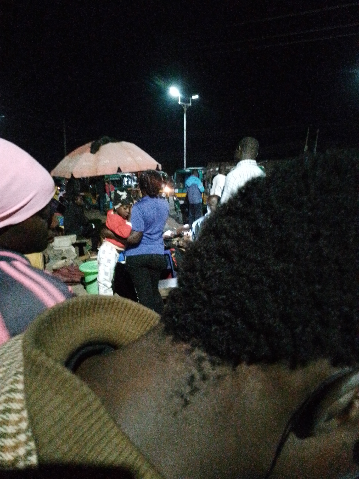 The image depicts a nighttime scene at a busy market in Bukuru, Nigeria. In the foreground, the back of a person wearing a dark, curly wig and a green sweater is visible. To the left, a person wearing a pink headscarf and a striped shirt is partially visible. In the mid-ground, a woman in a blue shirt holds a child wearing a red sweater and white pants. They are standing near a large, tan umbrella and several market stalls. Other individuals are scattered throughout the market, with some illuminated by a streetlamp in the background. The environment appears to be an outdoor market with temporary structures and an unpaved ground surface.