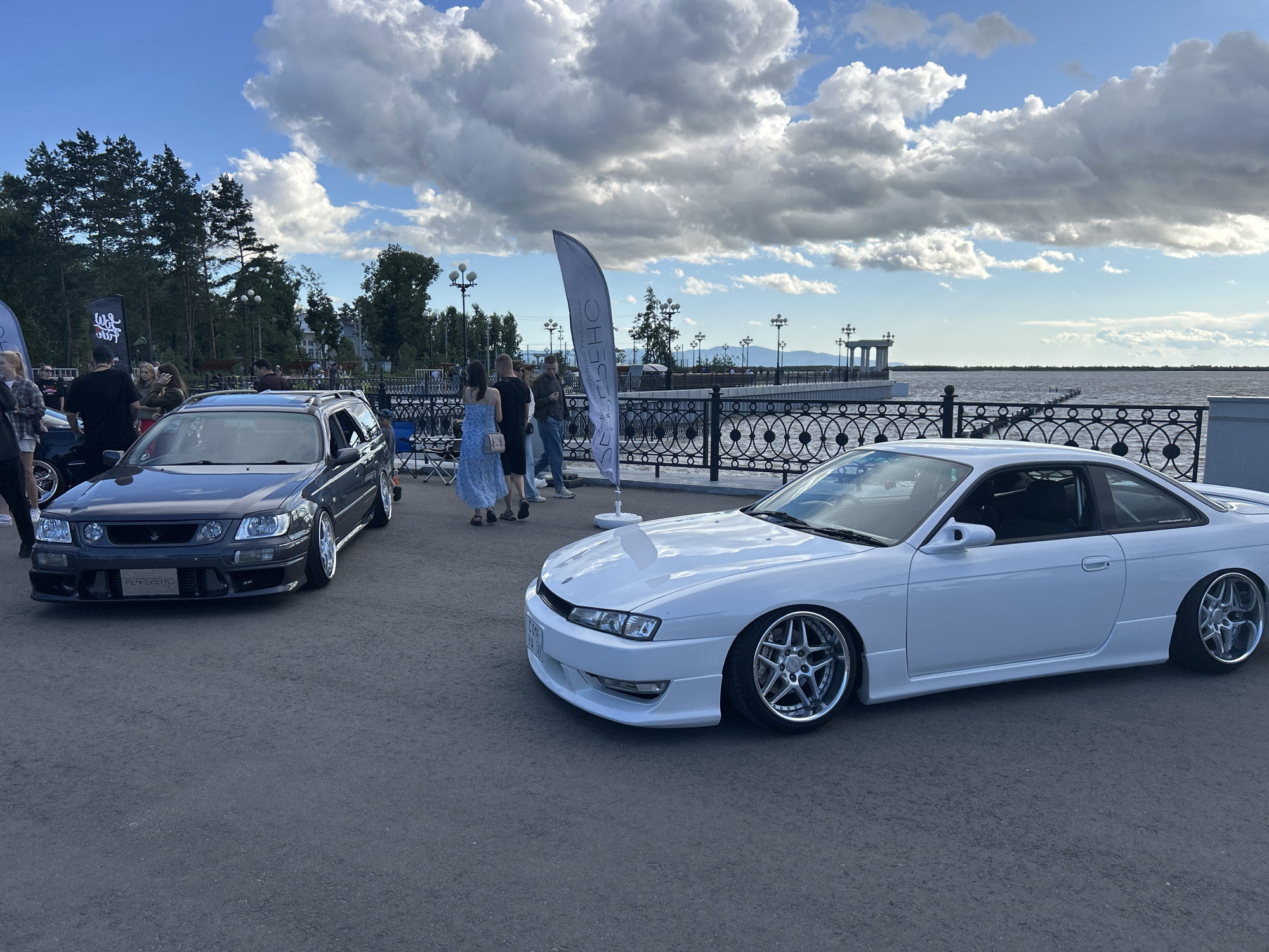 The image captures an outdoor car show event likely held in Khabarovsk, Russia, given the presence of Russian text on a banner. In the foreground, a white coupe and a dark-colored station wagon are prominently displayed. The white coupe, likely a Nissan Silvia S14, is positioned on the right side of the frame, showcasing its lowered stance and custom rims. The station wagon, appearing to be a Nissan Stagea, is on the left, also modified with aftermarket wheels. A group of people, dressed casually, are mingling around the cars, some appearing to be spectators and others potentially participants or organizers.
In the background, a body of water, likely the Amur River, stretches across the horizon, bordered by a promenade with a decorative railing and lampposts. A tall, white banner with Cyrillic lettering, reading "OTSH" and "PERHO", stands between the two cars, hinting at the event's name or sponsors. Beyond the promenade, distant hills and structures are visible under a partly cloudy sky. The overall atmosphere suggests a sunny day, possibly late afternoon, given the lighting conditions. The ground is paved, consistent with a city park or waterfront area.
