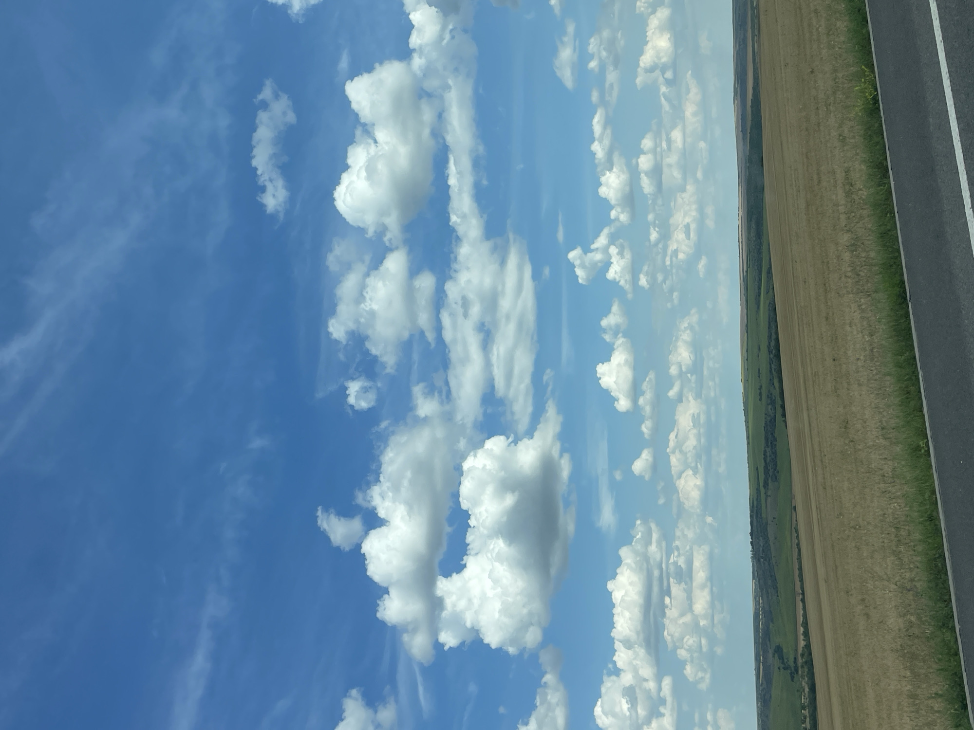 The image depicts a vast, rolling landscape under a bright blue sky dotted with fluffy white clouds. In the foreground, a dark asphalt road with a white stripe runs horizontally. Beyond the road, a field of dry, yellowish-brown grass extends into the distance. Further back, the terrain transitions to undulating green hills, some with patches of trees. The horizon is a soft line where the land meets the sky. The overall impression is one of a serene, rural environment, possibly captured from a moving vehicle on a clear day. This scene is representative of the countryside surrounding Comrat, the capital city of Gagauzia, an autonomous region of Moldova, characterized by its agricultural lands and gentle topography. There are no people or structures visible in the frame.