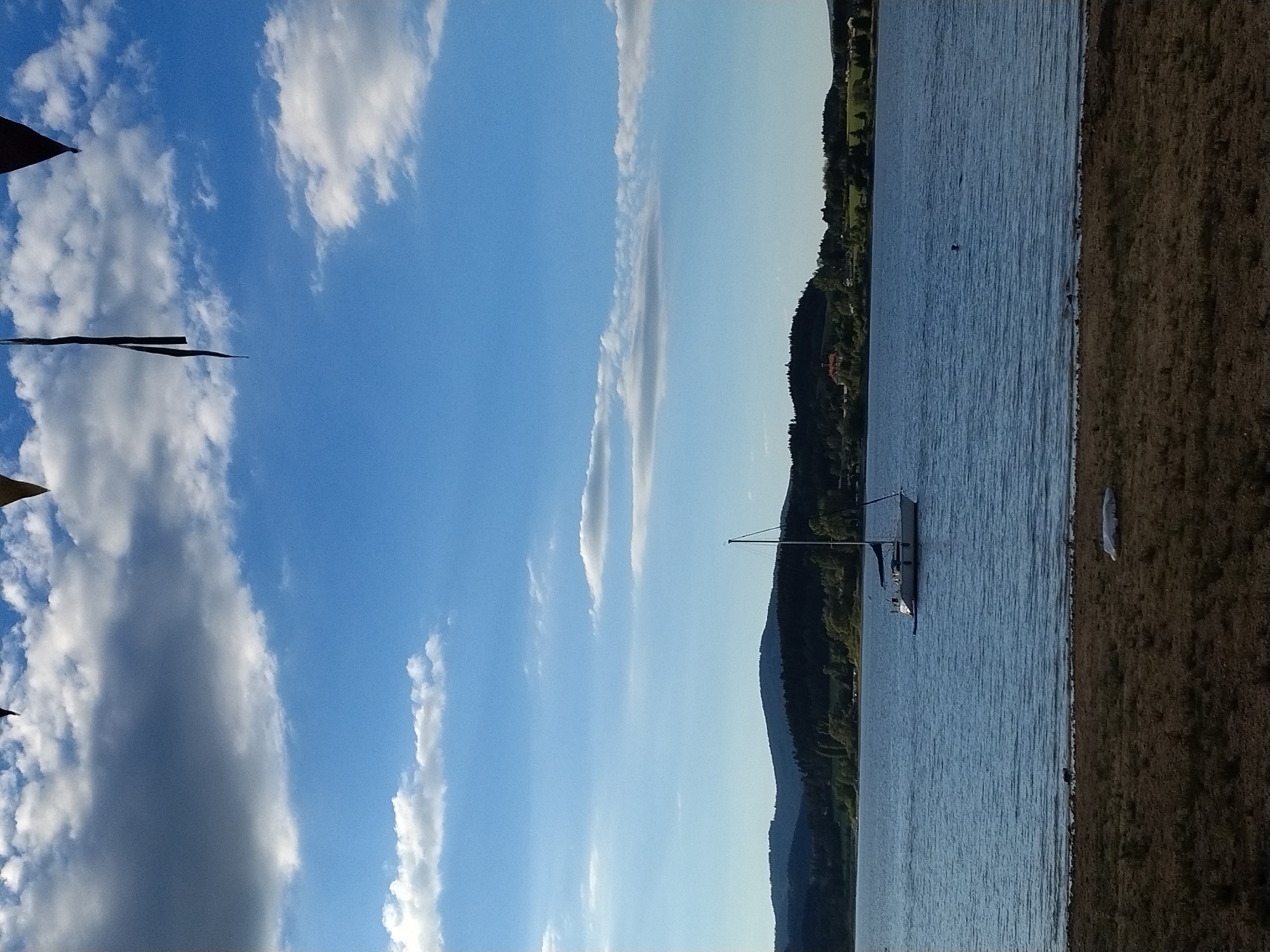 This is a vertical, eye-level, outdoor photograph captured on a sunny day, likely during the late afternoon given the lighting and cloud formations. The image depicts a tranquil scene at the Slapy Reservoir in Radslav, Czechia.
In the foreground, a dry, pebbly shore leads down to the calm, blue water of the reservoir. The water is rippled, suggesting a light breeze. There is a small, white object on the shore, possibly a piece of debris or a covered item.
The middle ground is dominated by the expansive reservoir. A white sailboat with a blue canopy is anchored or moored in the water, its mast reaching towards the sky. The boat appears to be unoccupied. Further in the distance, on the far side of the reservoir, is a dense, dark green treeline that forms a rolling hillside. Scattered amongst the trees are hints of buildings, suggesting a small settlement. The distant hills are bathed in a soft, muted light.
The sky above is a vibrant blue, with scattered white and gray clouds. Some of the clouds are cumulus, with soft, billowy shapes, while others are thinner and more wispy, stretched across the horizon. At the very top of the image, the dark shapes of triangular banners or flags are visible, dangling from an unseen structure above. These banners are a stark contrast to the bright sky and natural landscape.
The overall atmosphere of the image is one of peacefulness and serenity, with the natural beauty of the landscape and the solitary sailboat creating a sense of calm.