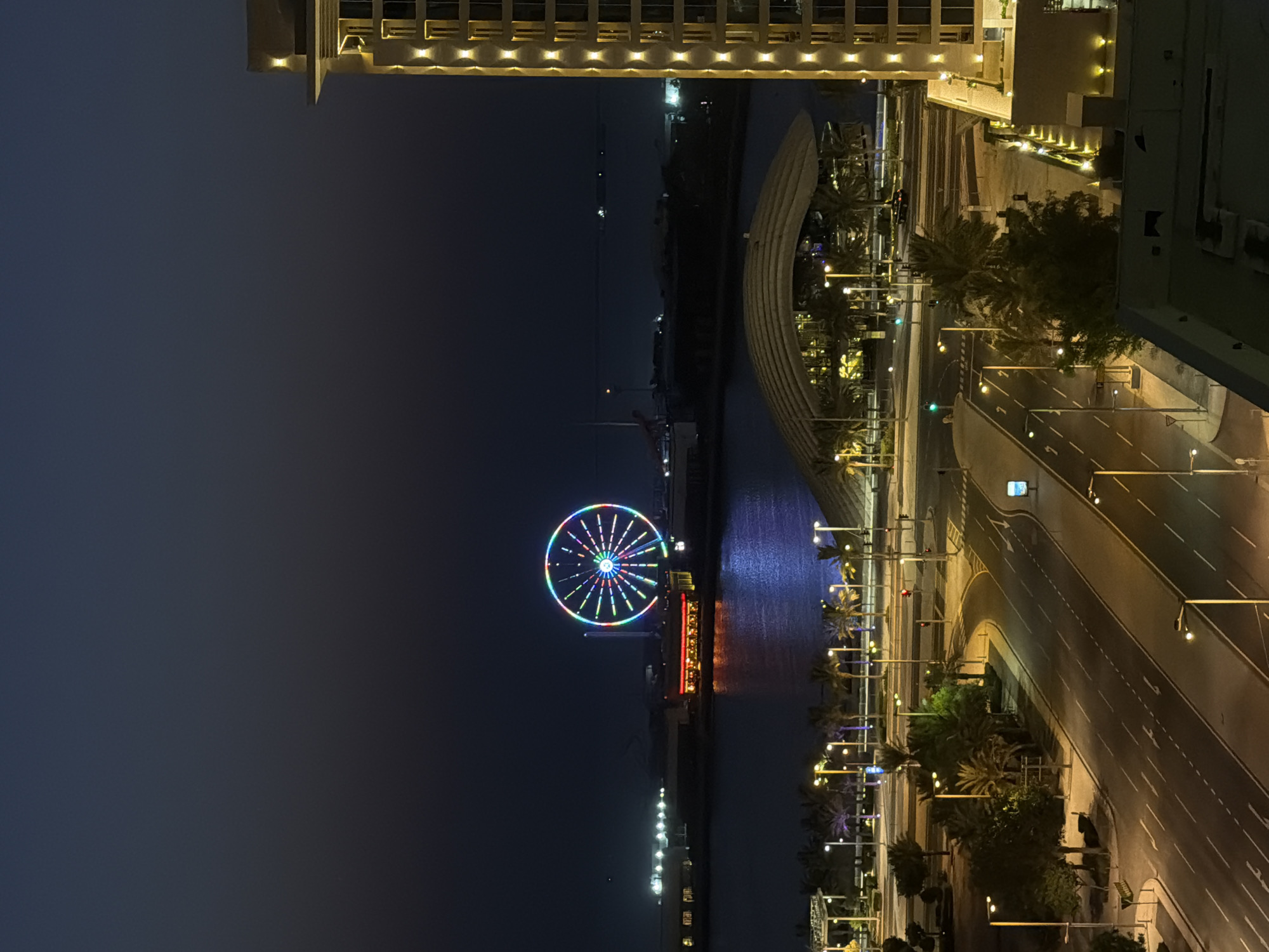 The image depicts a nighttime cityscape in Lusail, Qatar, featuring a brightly lit Ferris wheel in the distance across a body of water. The Ferris wheel displays a vibrant array of colors, illuminating the dark sky. In the foreground, a modern building with illuminated windows and accent lighting dominates the right side of the frame. Below the building, a multi-lane road with streetlights is visible, indicating urban infrastructure. Palm trees are scattered throughout the scene, contributing to the local atmosphere. The overall environment is dark, characteristic of nighttime, with artificial lights playing a significant role in illuminating the scene.