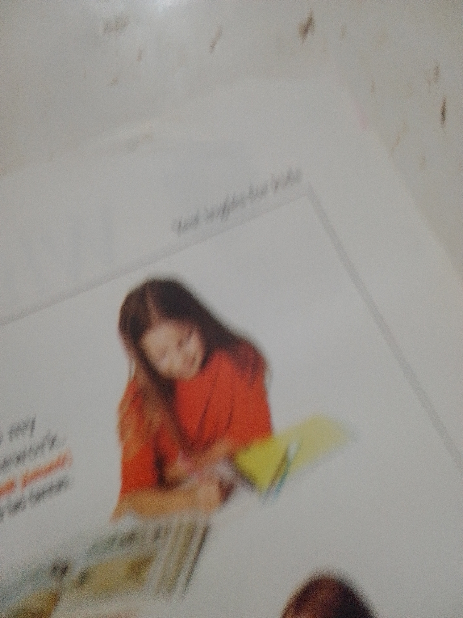 The image shows a blurry, top-down view of what appears to be a page from a book or magazine. A young girl wearing an orange shirt is visible, leaning over a book or papers on a surface. She is the primary subject. In the background, there is faint text that is mostly illegible due to the image's blurriness. The scene suggests an indoor setting, possibly a study area. There is no indication of a specific location in the city of La Tagua, Venezuela, or any contextual information that would place the image in that city. The image is not clear enough to provide further details about the environment, specific objects, or actions beyond the girl's posture.