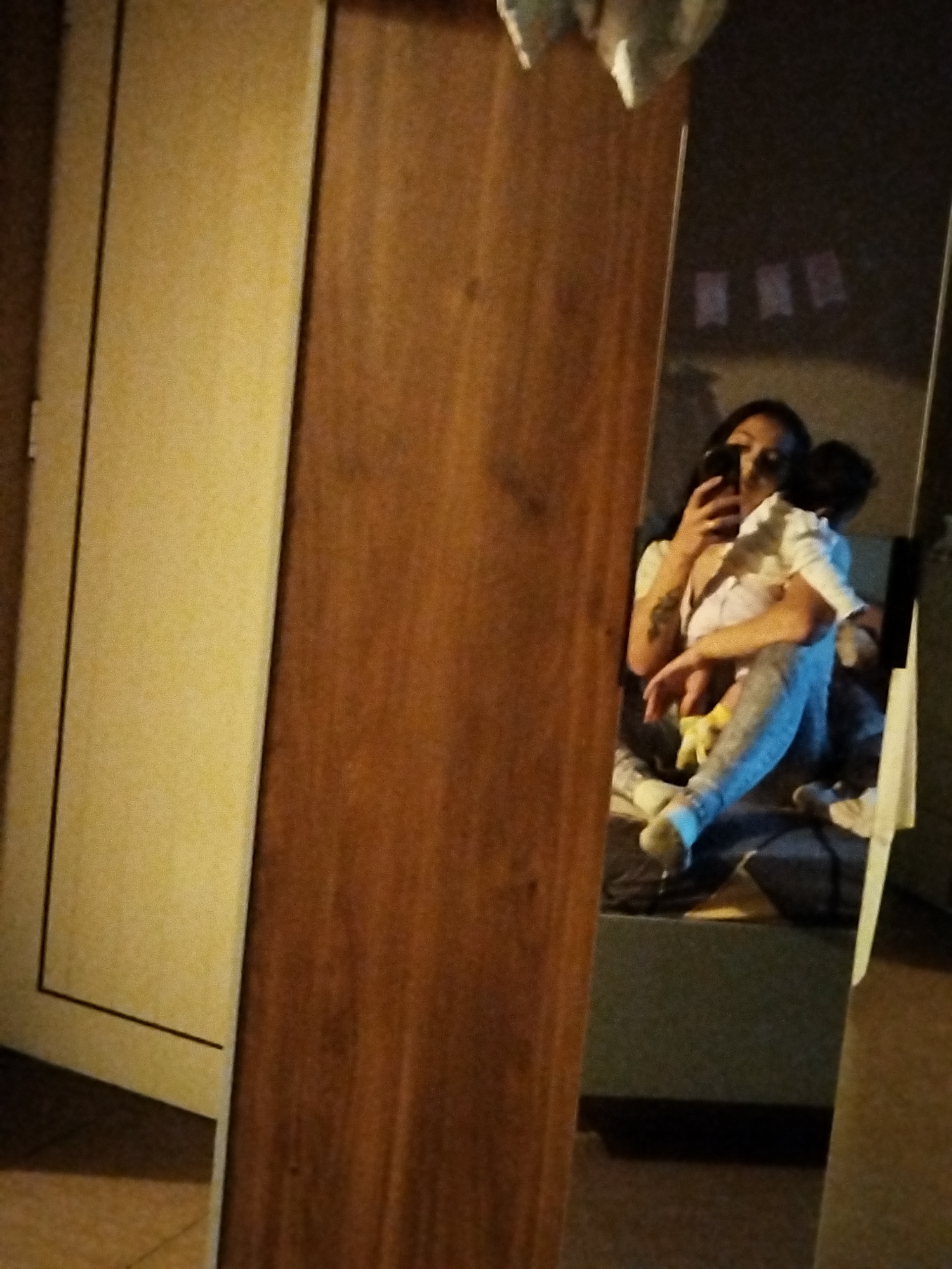The image is a reflection in a mirror, capturing a woman holding a child while taking a selfie with a phone. The woman is wearing light-colored clothing. The child is also wearing light-colored clothes. They are sitting on a surface or bed. The background is slightly blurred, but indistinct objects are visible. A wardrobe or cabinet is visible in the left foreground, obscuring part of the view. The location is unknown.