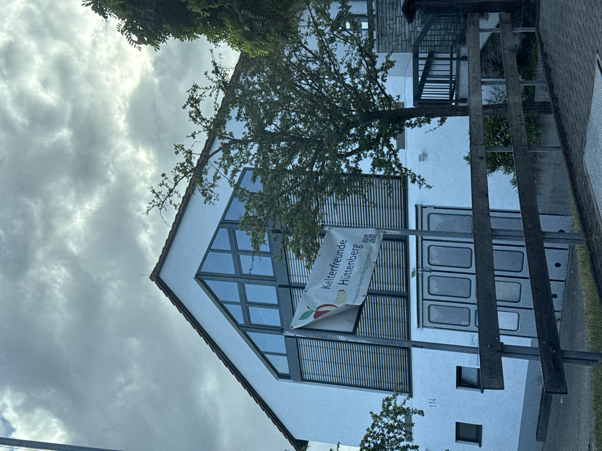 This media file shows the exterior of a white building with a triangular roof. A large tree with green leaves partially obscures the front of the building. A banner is hung on the building, featuring an apple graphic and text. The text on the banner reads "Kelterfreunde Hüttenberg" and "kelterfreunde-huettenberg.de". There are also QR codes visible on the banner. The building has several windows, some with dark blinds. To the right of the building, there is a fence or railing and some greenery. The sky is cloudy and overcast. A house number "114" is visible on the building. The scene appears to be a daytime shot in Volpertshausen, Germany. The overall mood is neutral, with no strong emotional cues conveyed.