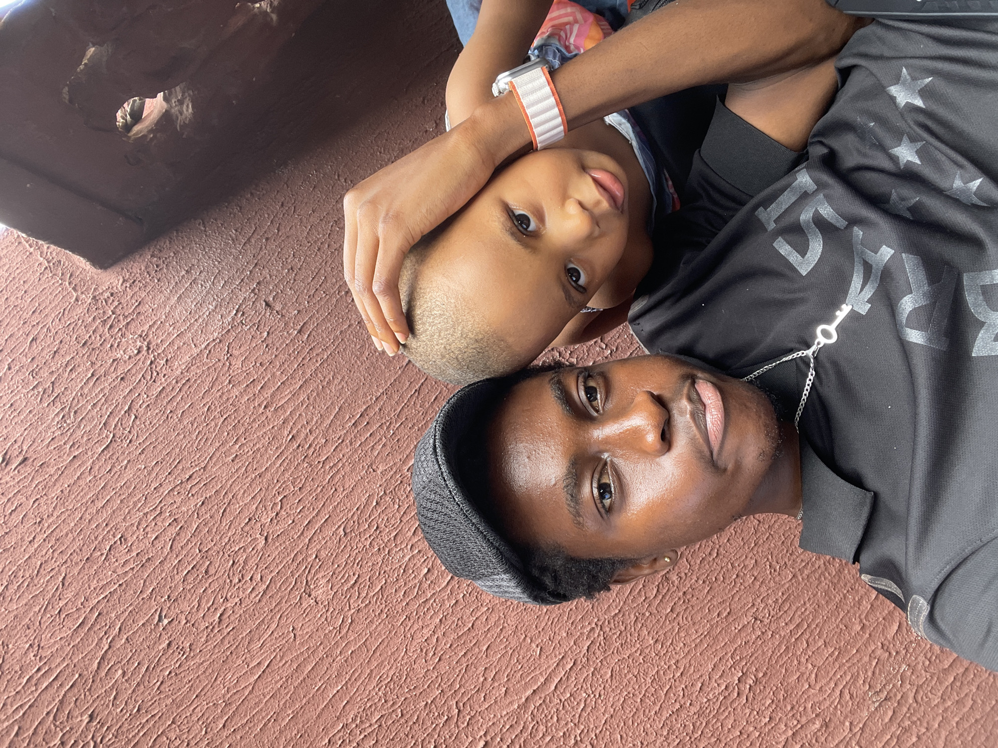 This daytime image captures an adult male and a child lying side-by-side on a textured, reddish-brown surface, likely a wall or ground, and looking directly towards the camera. The scene, potentially in Onitsha, Nigeria, shows a dark, recessed architectural feature with a small hole in the upper left background, casting a shadow.
The adult male, positioned in the lower half of the frame, wears a dark grey beanie and a black short-sleeved t-shirt. His shirt is adorned with white stars and the visible text "EST. 1974". A silver chain necklace with a key pendant hangs around his neck. He maintains a neutral expression.
Next to him, the child, with very short hair, has one arm wrapped around the adult's head. The child is wearing a smartwatch with a distinctive white and orange striped band on their left wrist and a patterned top. The child also looks directly at the camera with wide eyes and a slightly parted mouth, conveying a calm demeanor. The image portrays a posed or candid moment of connection between the two subjects.