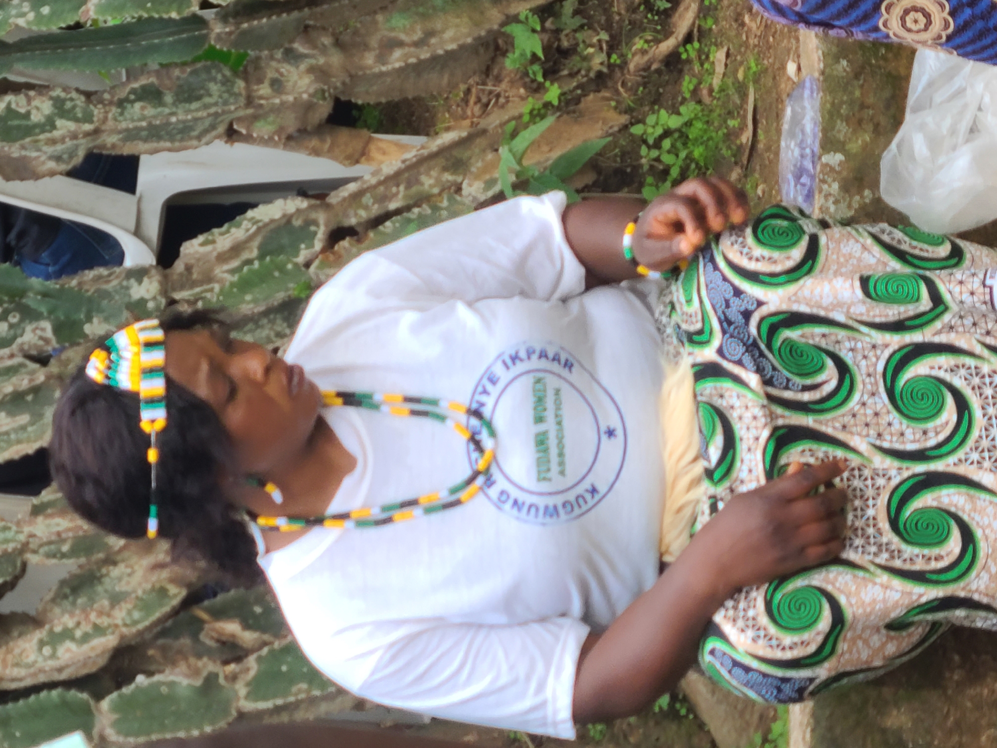 The image shows a woman wearing a white t-shirt with a circular logo and a colorful beaded headdress and necklace. She is seated outdoors, with a backdrop of large, segmented cacti. The woman's t-shirt has text that reads "NYE IKPAAR FEDANI WOMEN ASSOCIATION KUGWUNG". She is also wearing a bracelet with similar colors to her headdress and necklace. Her patterned wrap skirt, featuring swirls of green and black on a beige background, is visible. Her left hand is resting on her lap, and her right hand is touching the skirt. The scene suggests a cultural gathering or event. The lighting indicates it is daytime. The presence of cacti and the mention of "Kugwung" (a potential location or community name) in Jos, Nigeria, provide geographical context. The woman's expression is serene.