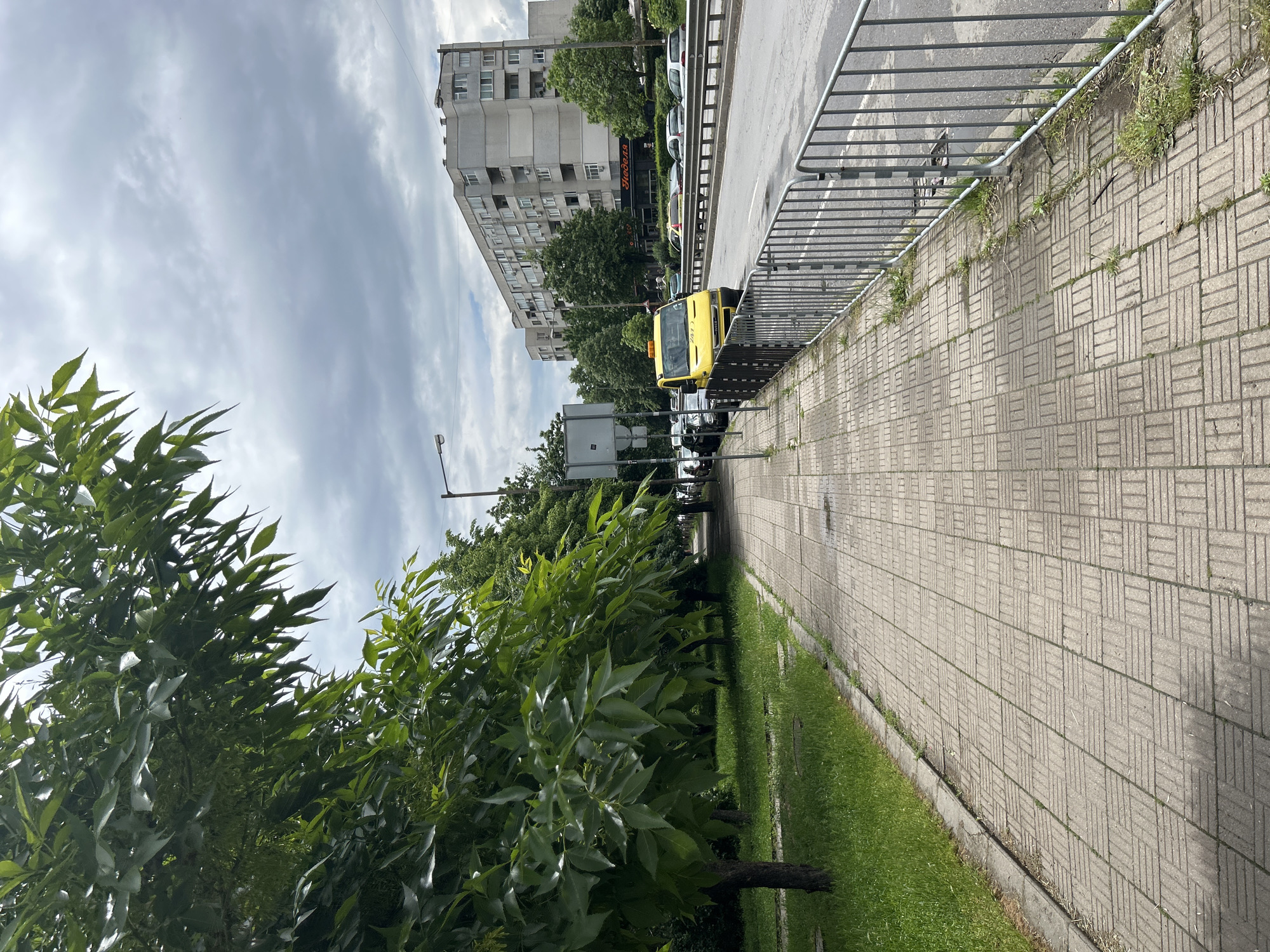 The image shows a sidewalk in a city, likely in Unknown, Unknown. The sidewalk is paved with gray rectangular blocks. To the right is a metal railing separating the sidewalk from the road. A yellow truck is parked alongside the road. Beyond the road is a multi-story building. The left side of the sidewalk is lined with green trees and grass. The sky is overcast.