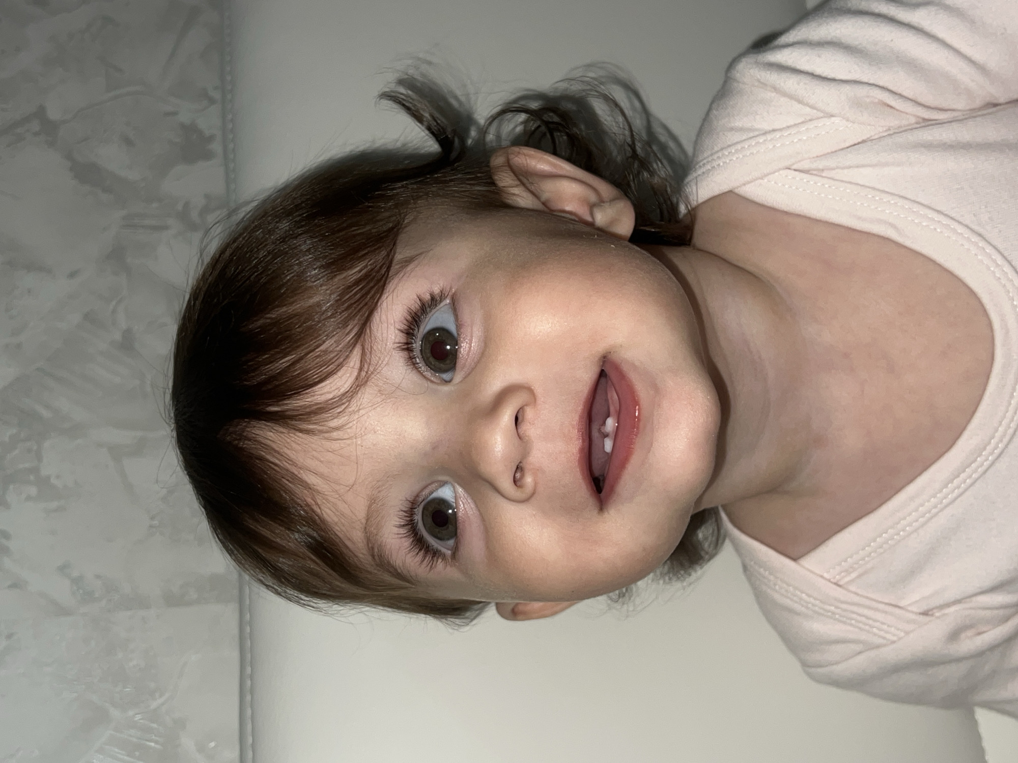 A close-up, slightly rotated, medium shot depicts a young child with brown hair and light skin. The child is wearing a light pink, long-sleeved bodysuit. The background appears to be a light-colored surface, potentially a bed or a changing table. The child's eyes are wide and brown, and the mouth is slightly open, revealing upper teeth. There is no indication of the location of Elena, Bulgaria.