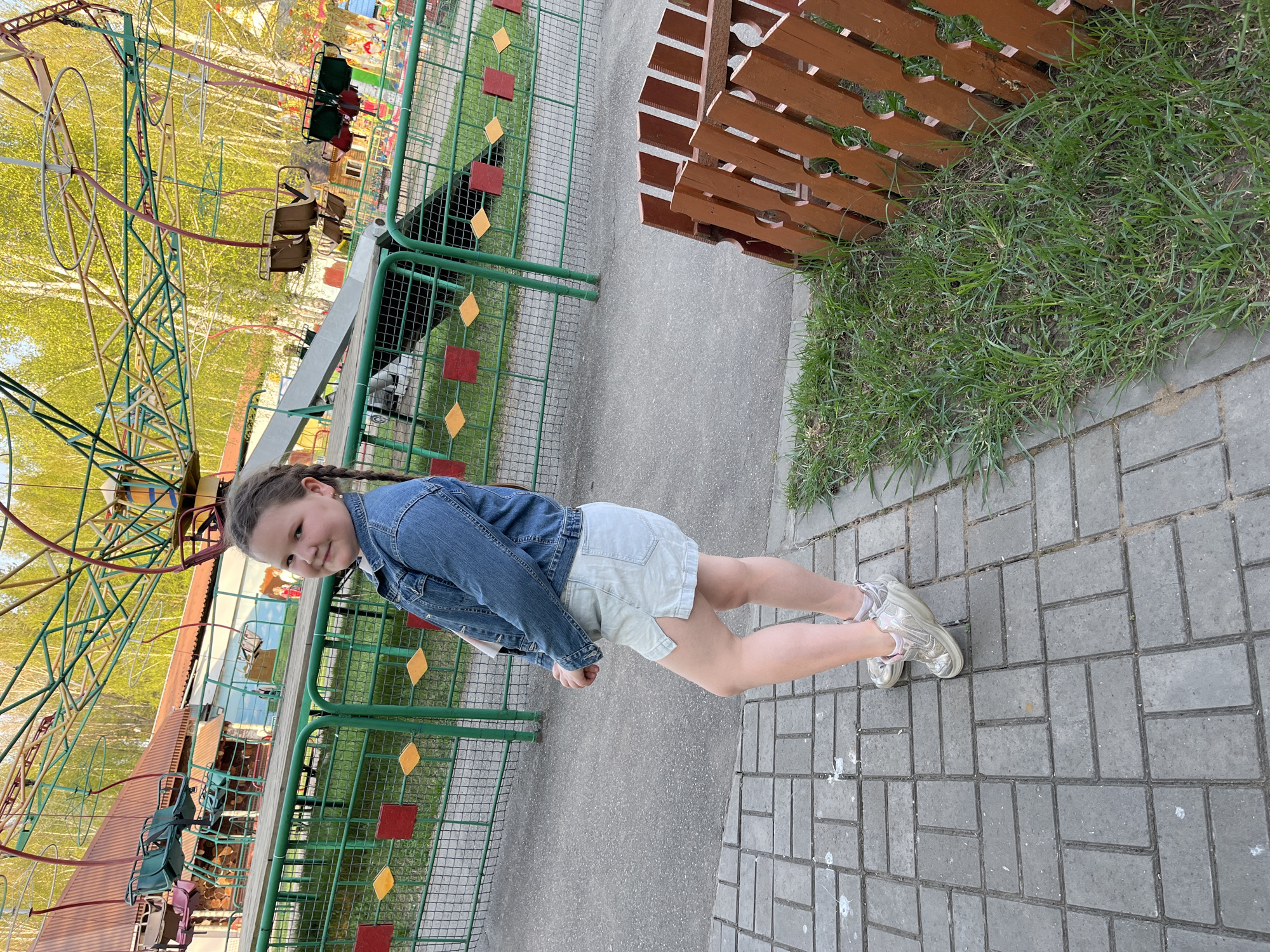 A young girl stands on a gray-bricked surface in what appears to be a park in Chaikovsky, Russia. She is dressed in a denim jacket and light-colored shorts, with her hair styled in braids. Behind her, a green metal fence with decorative red and yellow squares lines a paved pathway. To the girl's left is a carousel-type amusement ride, with empty seats visible. The background includes trees with green foliage, suggesting it's spring or summer. A wooden fence is visible on the right.