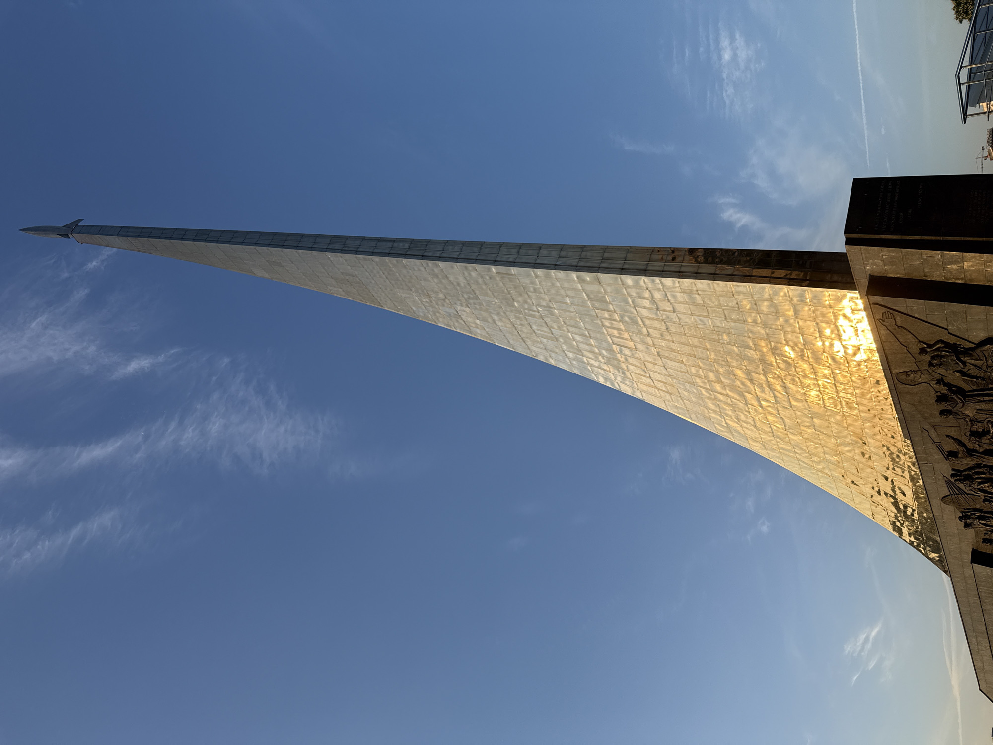 The image shows the Monument to the Conquerors of Space in Moscow, Russia. The monument is a tall, metallic structure shaped like a rocket rising into the sky. It is partly covered with reflective panels. The base of the monument is a darker, rectangular structure with bas-relief sculptures. The sky is blue with some wispy clouds. The sun reflects strongly off of the side of the monument.