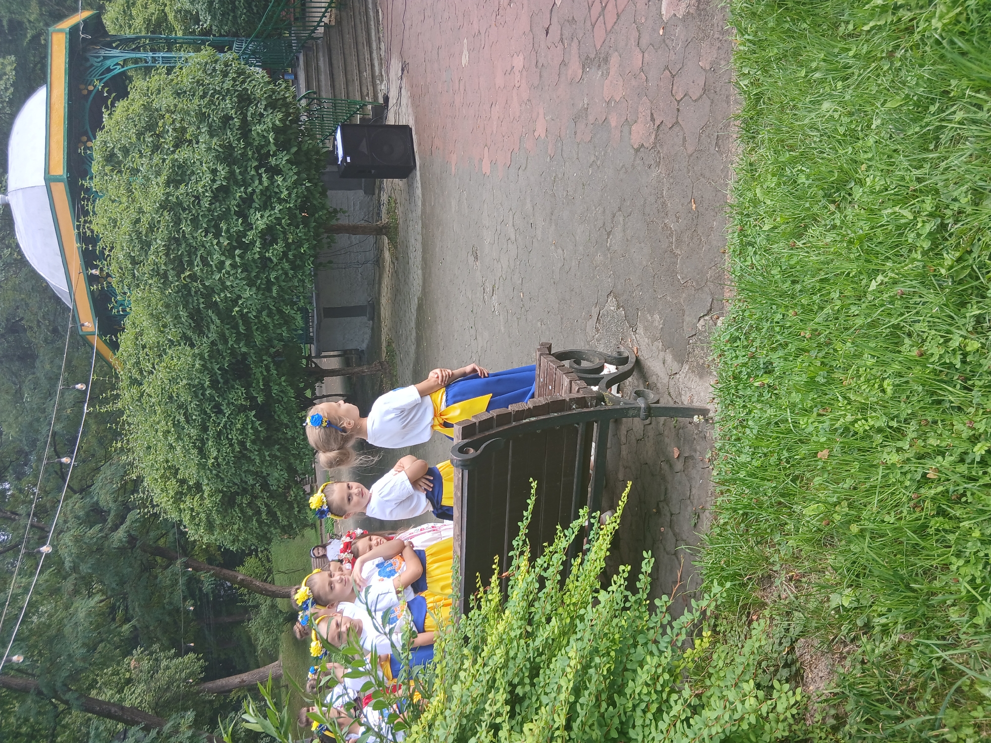 The image captures an outdoor scene in what appears to be a park or public square. In the foreground, a bench is visible, partially obscured by lush green foliage. Behind the bench, a group of young girls, dressed in traditional Ukrainian attire featuring blue and yellow elements, are standing. They appear to be looking in the same direction, suggesting they are observing something or waiting for an event. Further in the background, a decorative gazebo with a yellow and green painted trim stands prominently, and a set of stairs leads up to it. A large black speaker is placed on the paved ground near the stairs. Trees and greenery form a natural backdrop to the scene. The overall impression is of a community gathering or performance. While the image itself does not explicitly indicate Truskavets, Ukraine, the traditional attire of the children strongly suggests a Ukrainian context, and such park-like settings with gazebos are common in Ukrainian cities.