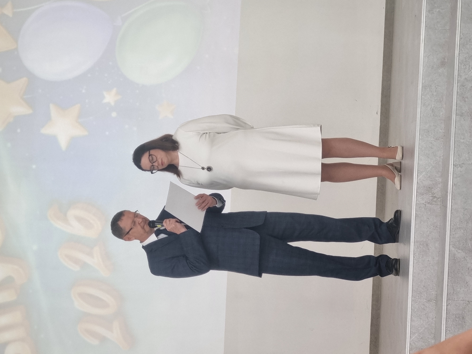 The image shows a man and a woman standing on a stage in front of a projected backdrop. The man, wearing a dark suit and glasses, is holding a microphone and a piece of paper, appearing to be speaking or reading. The woman, in a white dress and glasses, stands beside him with her hands behind her back. The backdrop features large, stylized numerals "2026" and images of balloons and stars. The setting suggests an event or ceremony. No specific location in the city of Chaikovsky, Russia can be identified from the image.