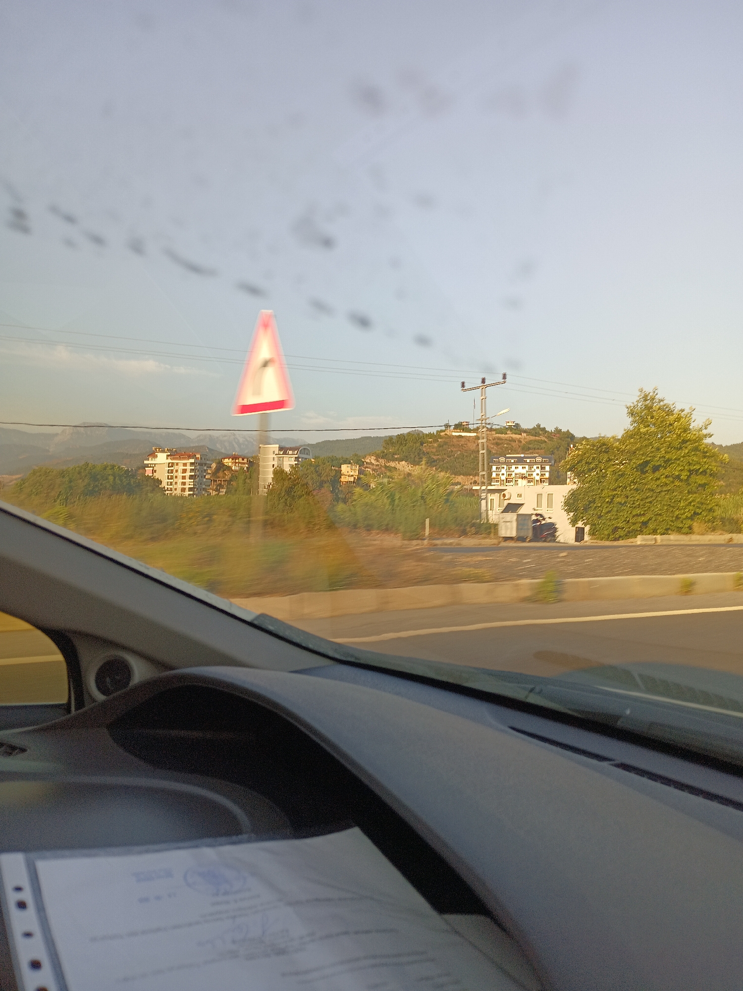 The image is a point-of-view shot from inside a vehicle, looking out through the windshield. The view is slightly blurred due to motion and possibly smudges on the glass. The foreground shows the dashboard and a document with some writing and a seal.
The middle ground reveals a landscape with trees and buildings. A prominent triangular traffic sign with a red border and white interior, indicating a curve, is visible. Beyond the trees, several multi-story buildings, likely apartments, are situated on a hillside. A power line pylon and overhead wires are also present. In the distance, mountains can be seen under a clear sky, suggesting an outdoor, likely rural or semi-rural, environment. The lighting suggests it is daytime, possibly late afternoon due to the quality of the light. The presence of multi-story residential buildings and the overall landscape are consistent with the outskirts or less densely populated areas of Alanya, Turkey, which is known for its hilly terrain and coastal views.