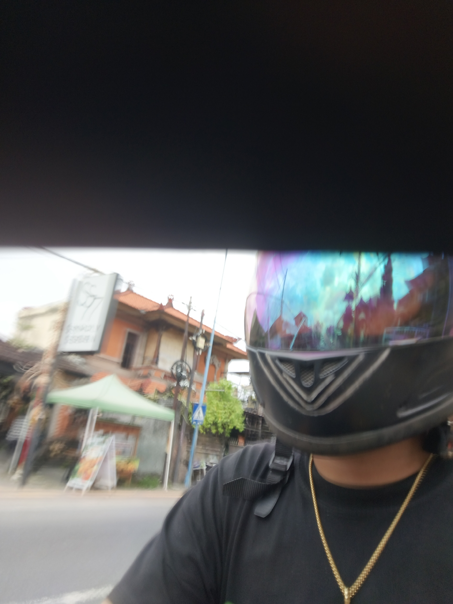 A selfie taken from the perspective of a person wearing a black t-shirt and a black motorcycle helmet with a reflective visor, in a city street scene. The visor reflects buildings, trees, and a cloudy sky. In the background, there's a building with an orange facade and a red tile roof, a green tent, a signpost, and a street. The image is blurry due to motion, suggesting it was taken while moving, likely on a motorcycle. The setting appears to be urban and tropical, consistent with a location in Denpasar, Indonesia.