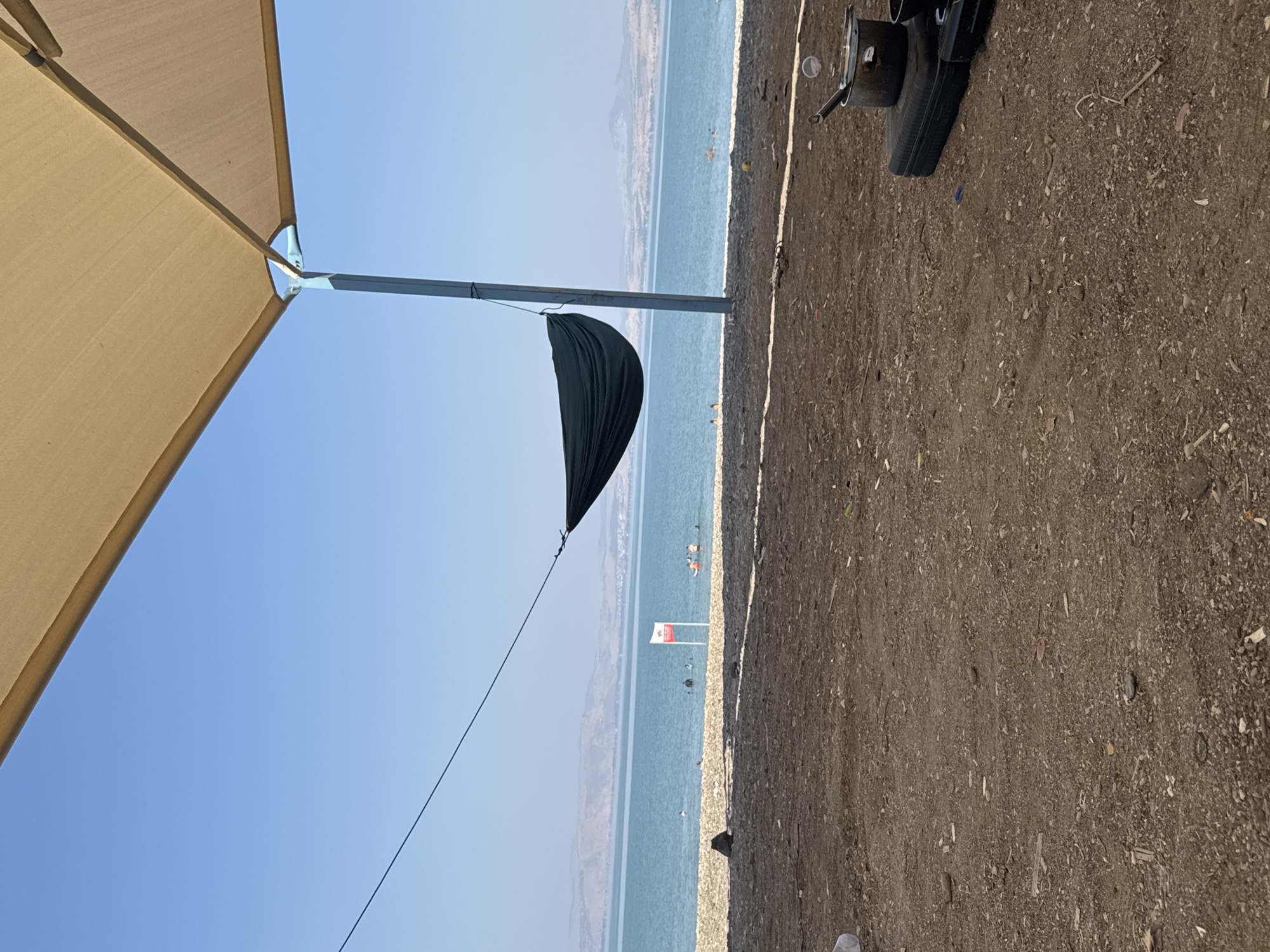 A dark green hammock is suspended from a tall pole and a taut wire, casting a shadow on the pebbly ground below. The hammock hangs over a beach area, with a calm, blue body of water in the background. A white and red flag is visible near the water's edge, with several people swimming and wading. In the distance, hills form a hazy horizon. In the foreground, on the right side of the image, a portable stove with a pot and lid is set up on a black case. The scene appears to be a campsite or picnic area, likely along the coast of the Sea of Galilee in the Golan Regional Council, Israel.