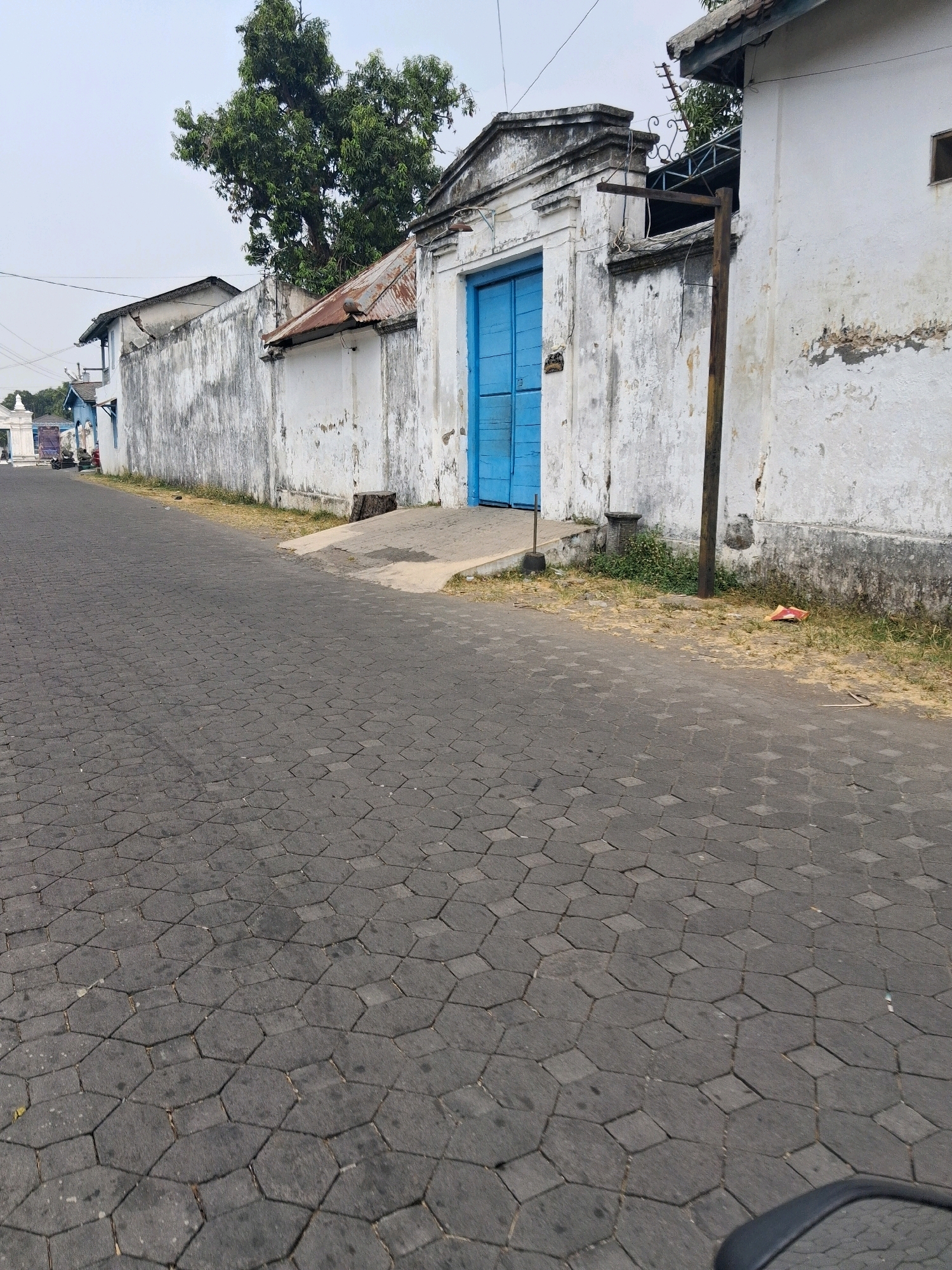 The image depicts a street scene in Surakarta, Indonesia. A dark, hexagonal-patterned cobblestone road occupies the foreground, leading towards a white wall with peeling paint. A large, bright blue double gate is set within this wall, framed by decorative architectural elements. A rusted metal roof extends from the top of the gate area, and a tall, thin wooden post stands next to the wall. Beyond the wall, a large tree with green foliage is visible, as are parts of buildings with white walls and darker roofs. The sky is overcast. The scene appears to be a quiet, possibly historic, part of the city.