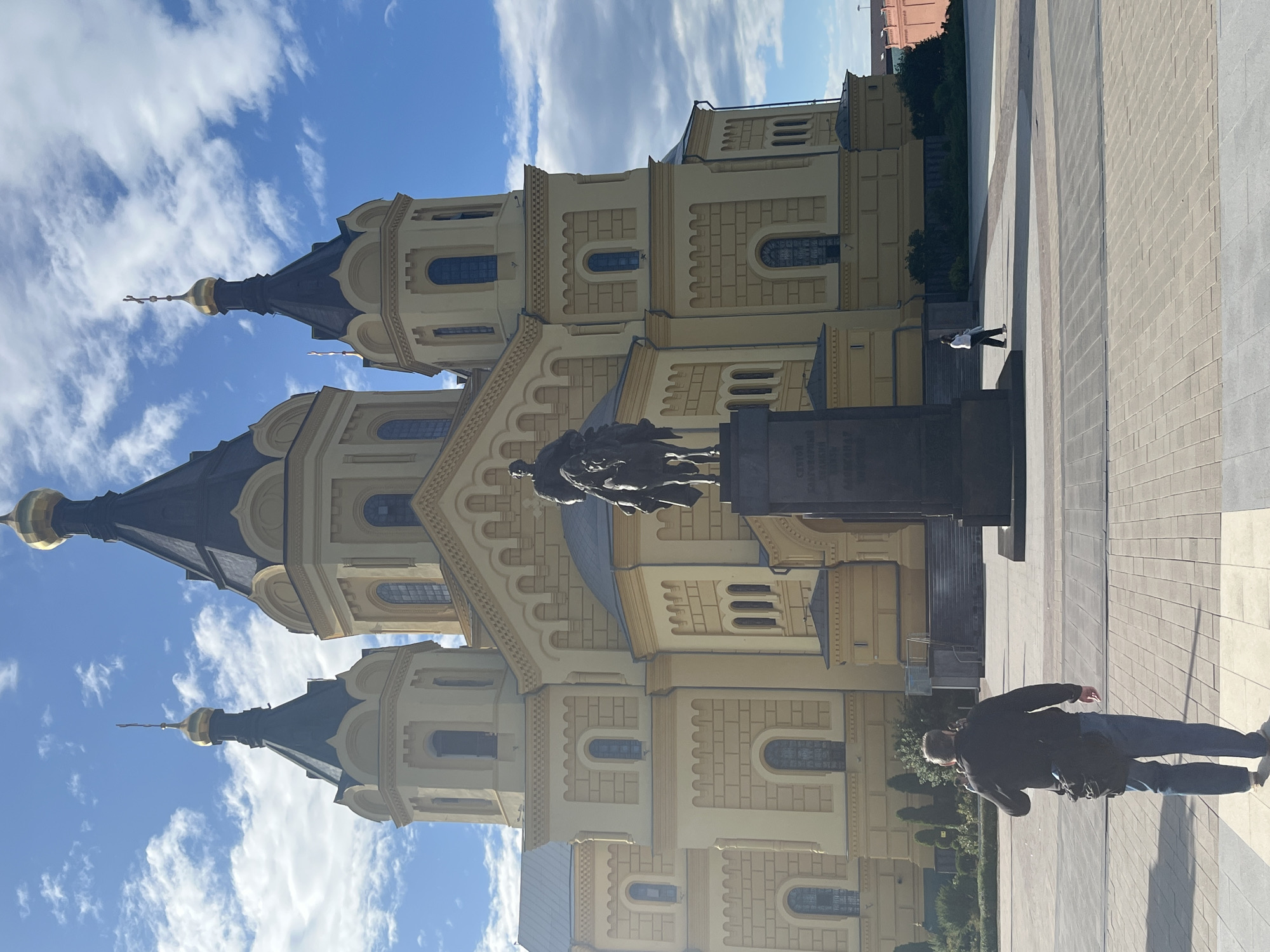 A low-angle, full-shot photograph captures a monument situated in front of a grand, yellow orthodox cathedral in Nizhny Novgorod, Russia. The cathedral, with its dark domes topped with golden crosses, dominates the background under a partly cloudy blue sky. In the foreground, a man wearing a black jacket and blue jeans walks away from the camera, carrying a black bag slung over his shoulder. To the right of the monument, a person in white walks away from the camera. The ground is paved with rectangular stone tiles. The monument itself is dark, with an equestrian statue at the top and an inscription on its base. The overall scene is bright and appears to be set during the daytime.