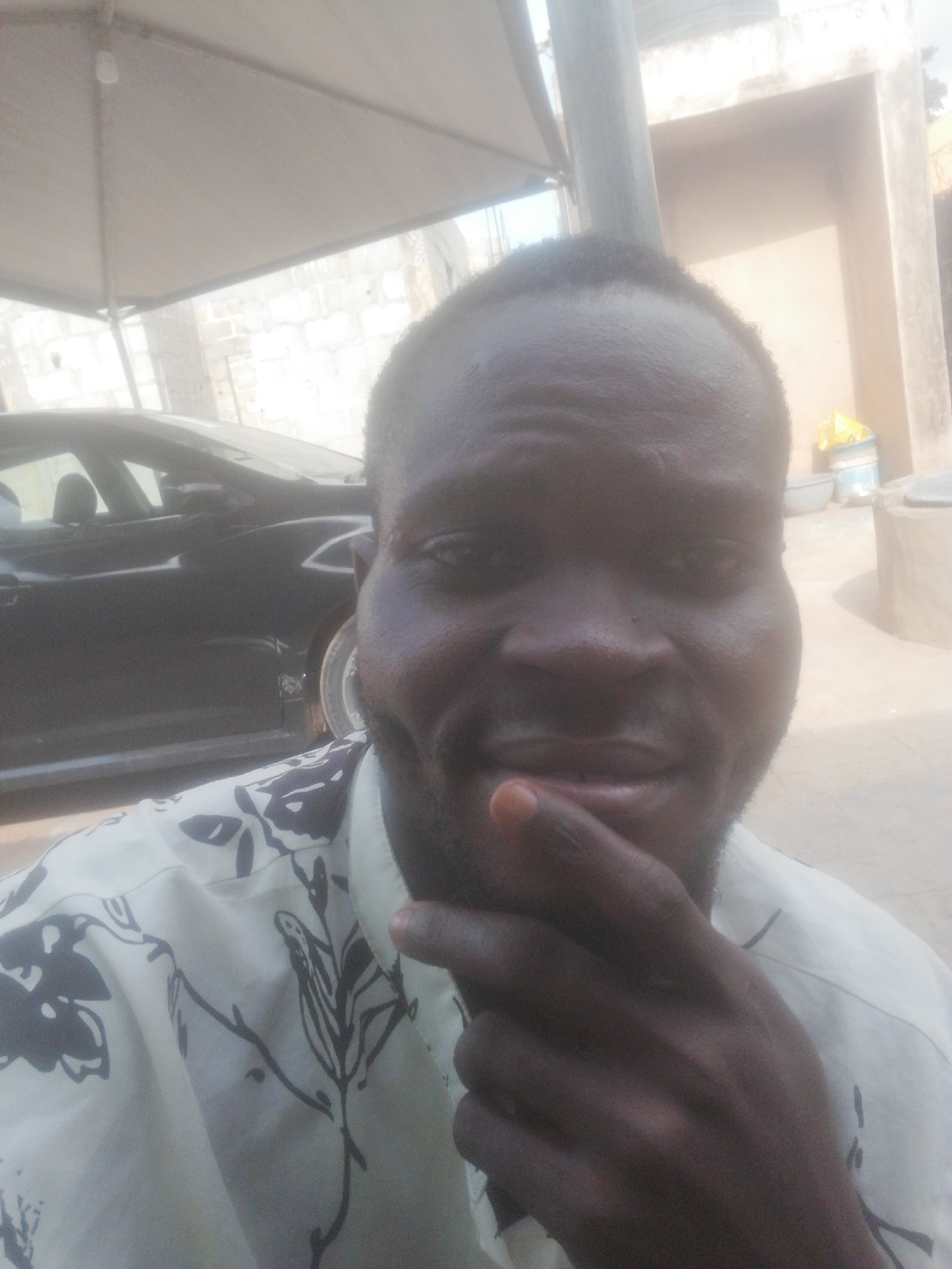 A man poses for a selfie. He is dark-skinned and wearing a floral print shirt. His finger is touching his chin. In the background, there is a black car under a carport-like structure, and a building or wall. The location is not discernible from the provided image and is labeled Unknown, Unknown.