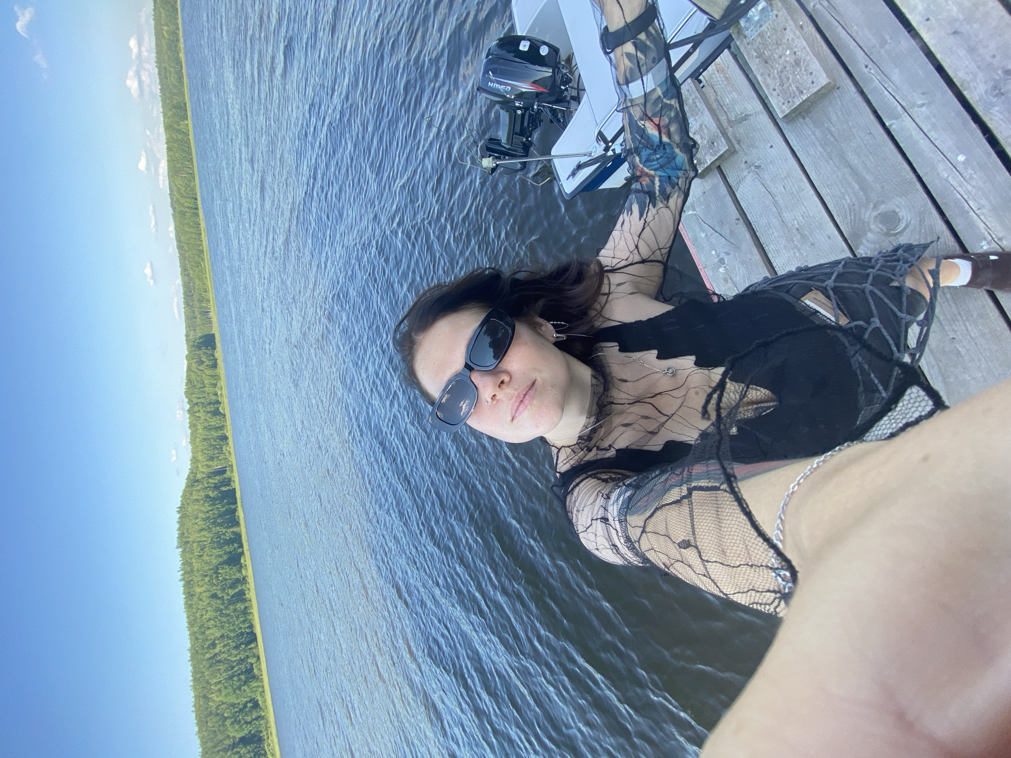A young woman stands on a wooden pier on the Kama River in Chaykovsky, Russia. She is wearing a black sheer top with an intricate pattern and a black bralette underneath. She also has on black sunglasses, a silver necklace, and a silver bracelet. Her left arm is visible, and she has tattoos on it. A small white boat with a black outboard motor is docked next to her on the pier. The river is calm, with gentle ripples, and a lush green forest lines the opposite bank of the river. The sky is a clear blue with a few scattered clouds.