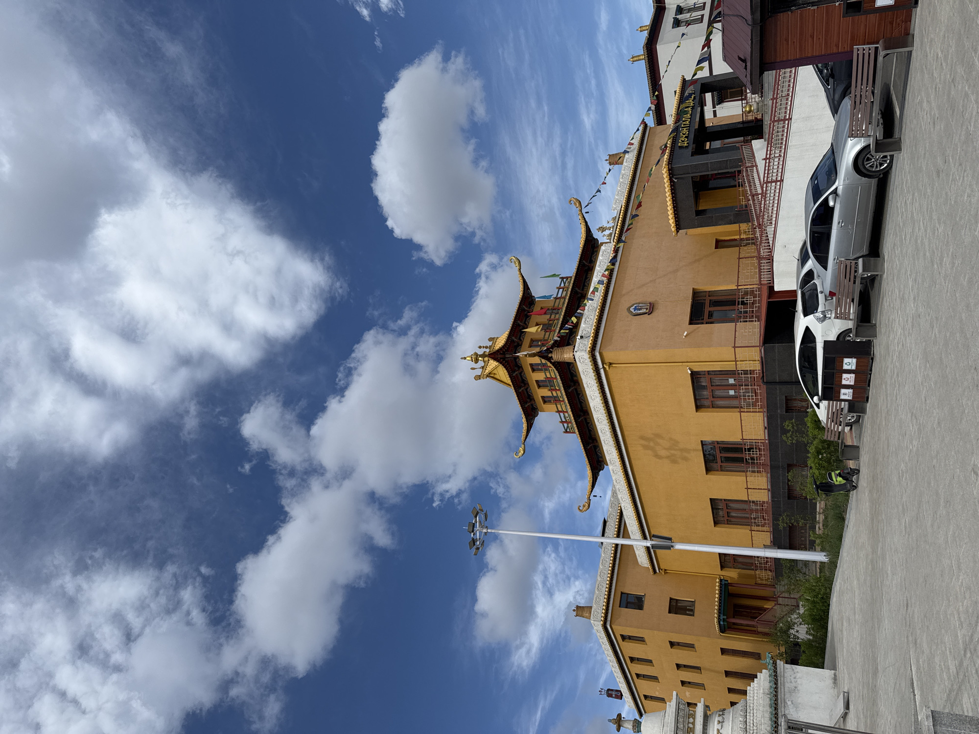 The image shows a multi-story building in Ulaanbaatar, Mongolia, set against a blue sky with scattered clouds. The building has a traditional architectural style with a predominantly yellow facade, a roof with ornate golden embellishments, and colorful prayer flags. A tall, white light pole is visible near the building. Parked adjacent to the building are a white and a silver car, with benches placed behind them. A paved sidewalk is visible in the foreground.
