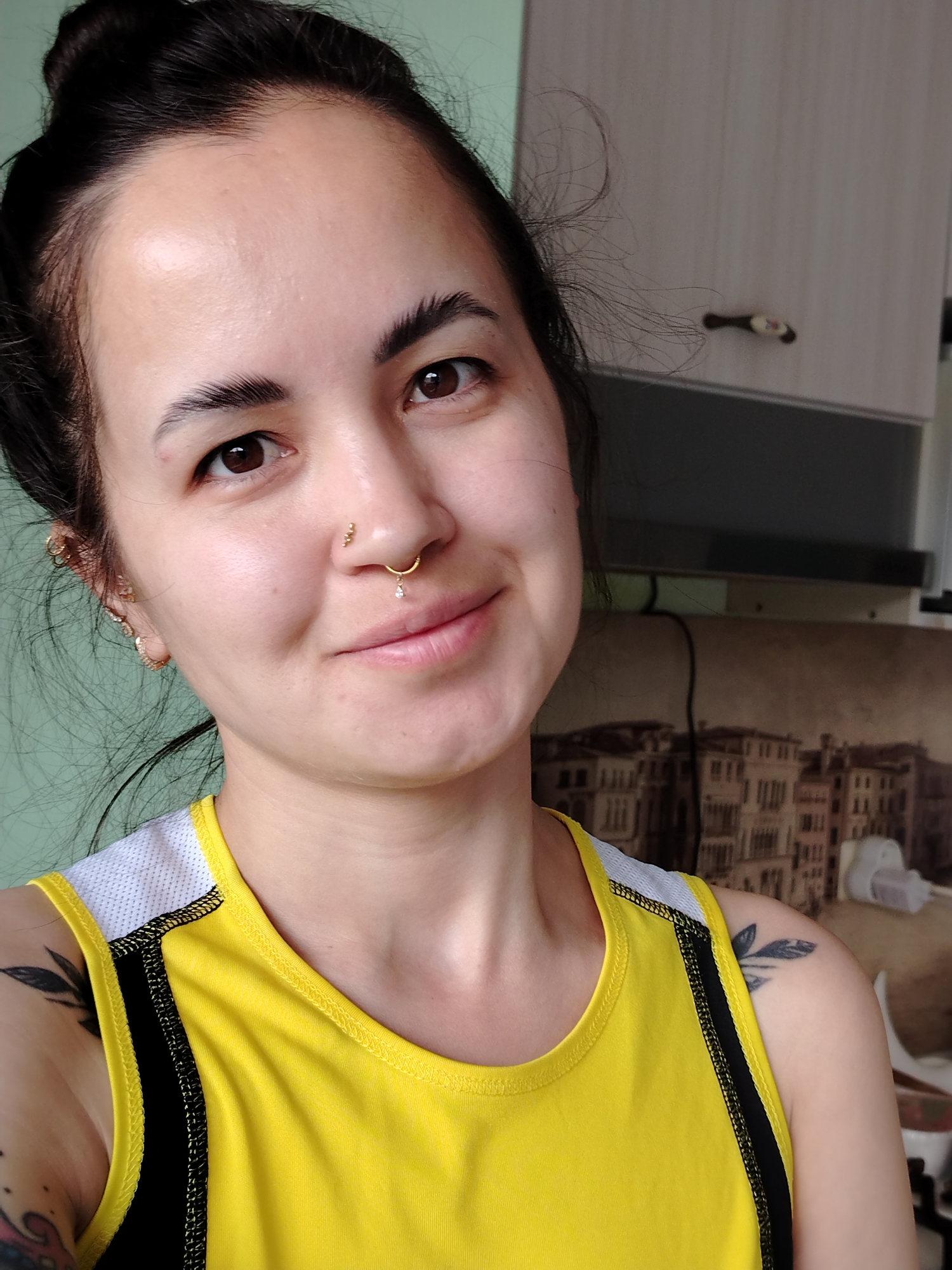 A woman is posing indoors. She has dark hair pulled back in a bun and wears a yellow and black athletic top. She has numerous piercings in her ear and nose. She smiles slightly. Tattoos are visible on her arms. The background features a light green wall and kitchen cabinets. A print of a cityscape hangs behind her. There is no information to confirm the location is Noviy, Russia.