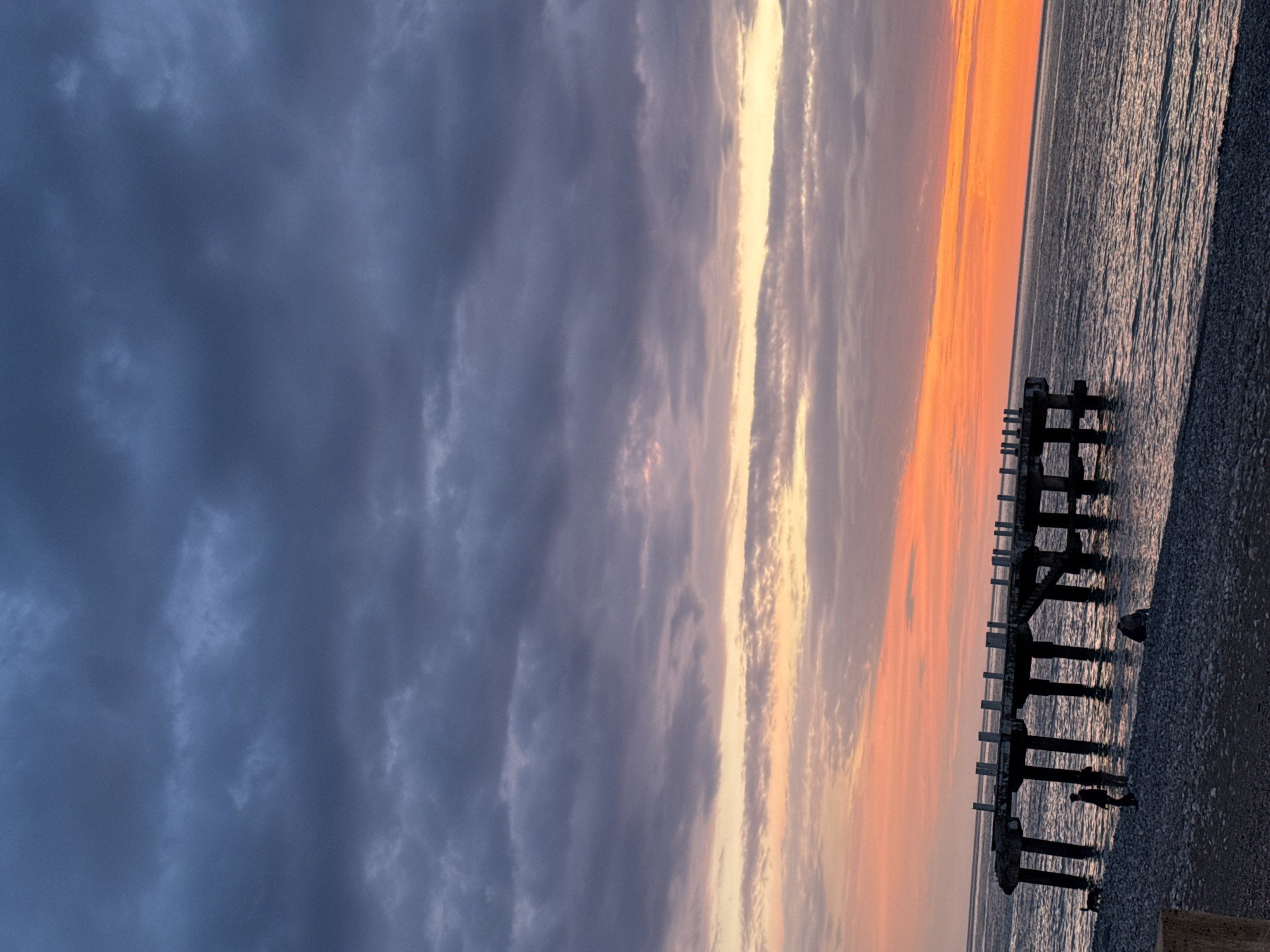 The image depicts a dramatic coastal scene at either sunrise or sunset, characterized by a striking interplay of light and shadow across the sky and water. The sky is bisected by varying light conditions; the left side is dominated by dense, dark blue-grey clouds, suggesting a heavy, overcast atmosphere or recent weather. Towards the right, the horizon is ablaze with vibrant orange and yellow light, indicating the sun's position. This bright light extends upwards, illuminating parts of the clouds in streaks of white, gold, and soft pinks, creating a dynamic contrast with the darker sky.
Below the horizon, a calm body of water, likely the sea or ocean, reflects the muted light, showing a textured surface with gentle ripples. Along the right side of the frame, a pebbled beach or shoreline extends, meeting the water.
A significant structural element in the mid-ground is a dark, silhouetted pier or jetty. This structure appears to be old or partially ruined, with several concrete pillars extending into the water and along the shore. Several small, silhouetted figures of people are visible near and under the pier. One figure is clearly standing beneath an archway of the structure, and another small shape, possibly a dog, is near one of the figures closer to the viewer. The overall atmosphere is serene yet dramatic, with the rich colors of the sky creating a powerful visual impact. There is no visible text in the image.
