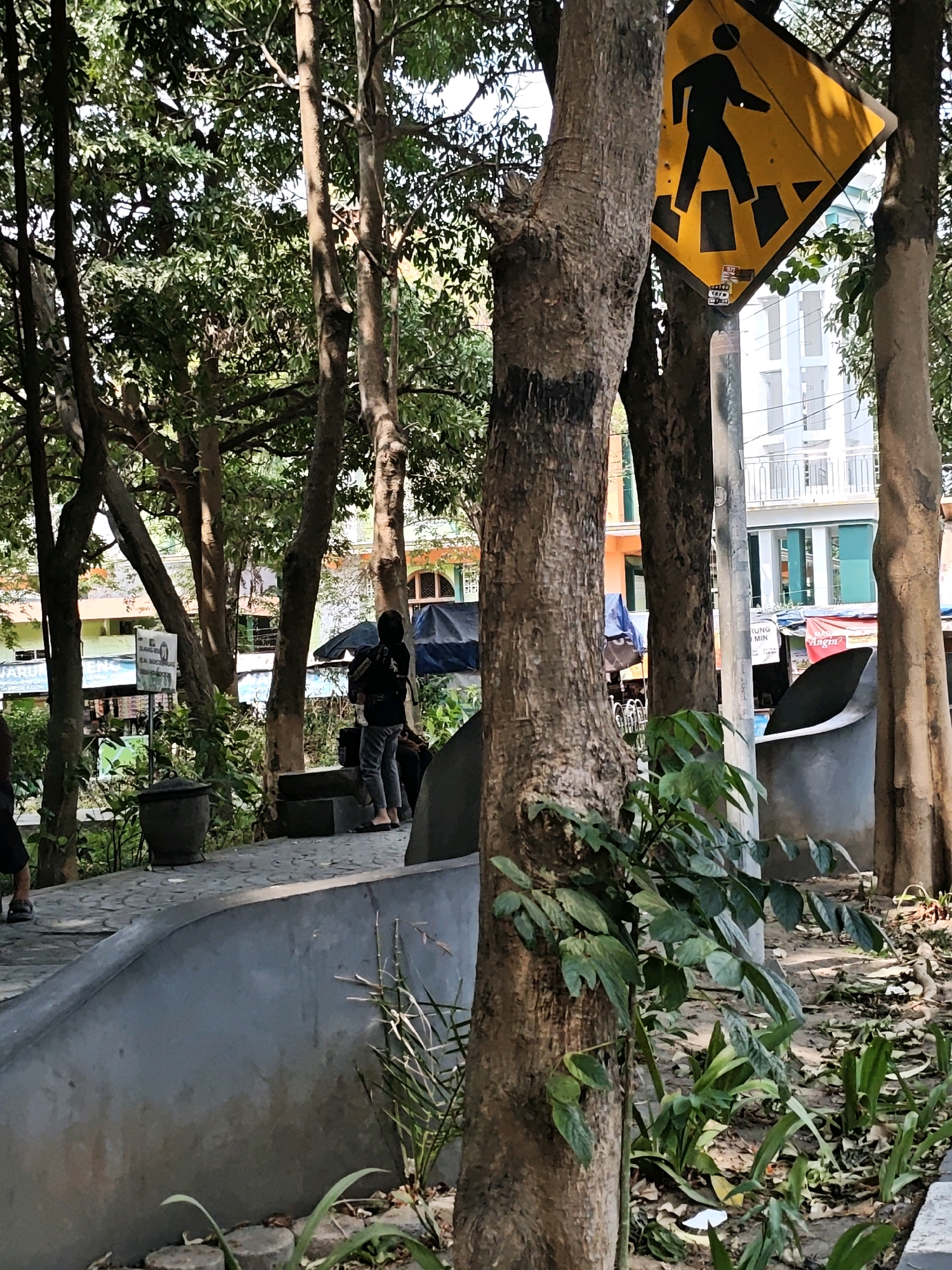 In Kediri City, Indonesia, a pedestrian crossing sign is visible amidst a park-like setting with trees and a pathway. A person wearing a dark hoodie and pants stands near the pathway, facing away from the camera. Buildings, some with balconies and windows, are visible in the background. The ground is paved with stones, and there are potted plants and loose leaves scattered around. The scene appears to be outdoors on a sunny day.