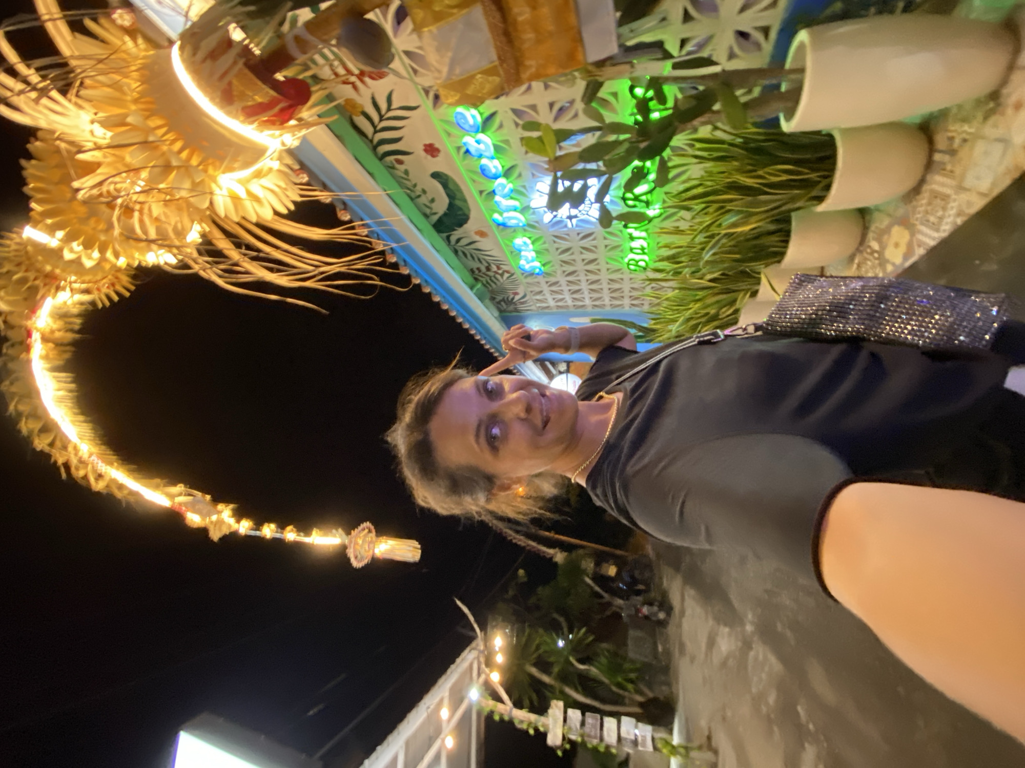 The image shows a woman taking a selfie at night in Nusa Penida, Indonesia. She is wearing a black t-shirt and a silver sequined clutch is visible. Behind her, there is a decorative structure with intricate carvings and bright yellow lights, resembling a dragon or a phoenix. The background features a wall with a tropical-themed mural, and several potted plants with long, green leaves. Neon blue lights spell out what appears to be "Bali" vertically on the wall. The woman is smiling and has her hand raised in a peace sign. The scene is illuminated by artificial lights, suggesting it is evening or night. The overall atmosphere is festive and vibrant, characteristic of a tourist destination.