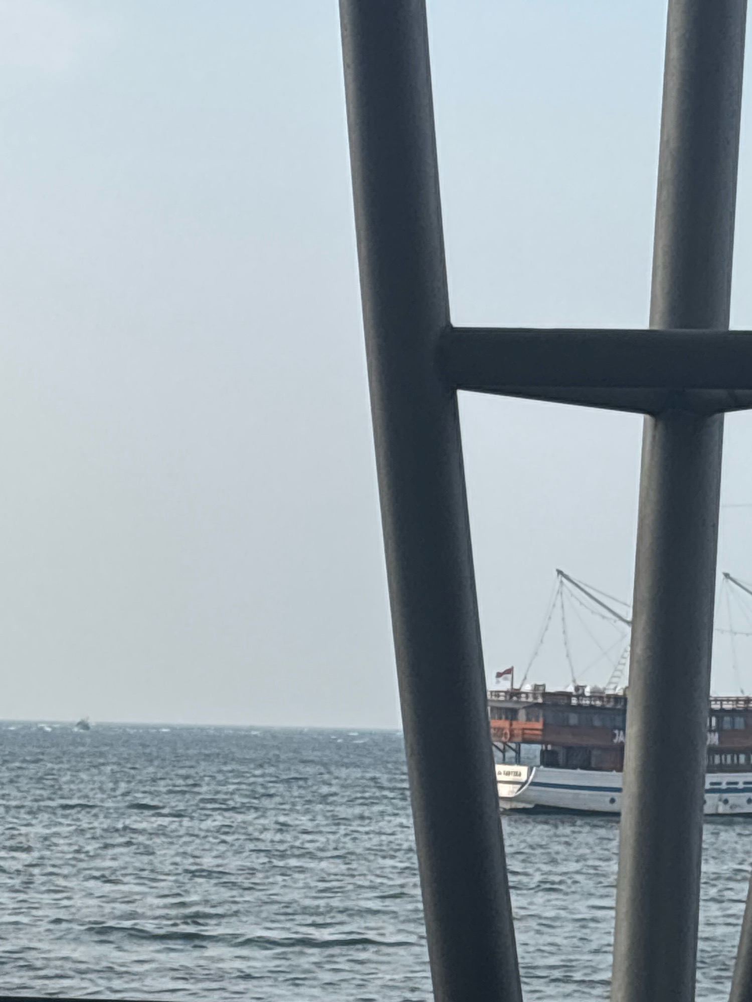 The image shows a view of the sea through a metal structure. On the water, there are two boats. The boat on the right is a large, multi-deck vessel with a reddish-brown upper section and a white hull. It has masts and rigging, suggesting it is a traditional sailing ship. A flag, possibly red and white, is visible on the upper deck. The boat on the left is much smaller, appearing as a speck on the horizon. The sea is a deep blue-grey with moderate waves. The sky is overcast with a pale, hazy light. The metal structure in the foreground consists of thick, dark grey cylindrical posts and horizontal beams, partially obscuring the view of the sea and boats. The scene is likely from a waterfront area in North Jakarta, Indonesia, given the presence of traditional Indonesian vessels and the maritime setting.