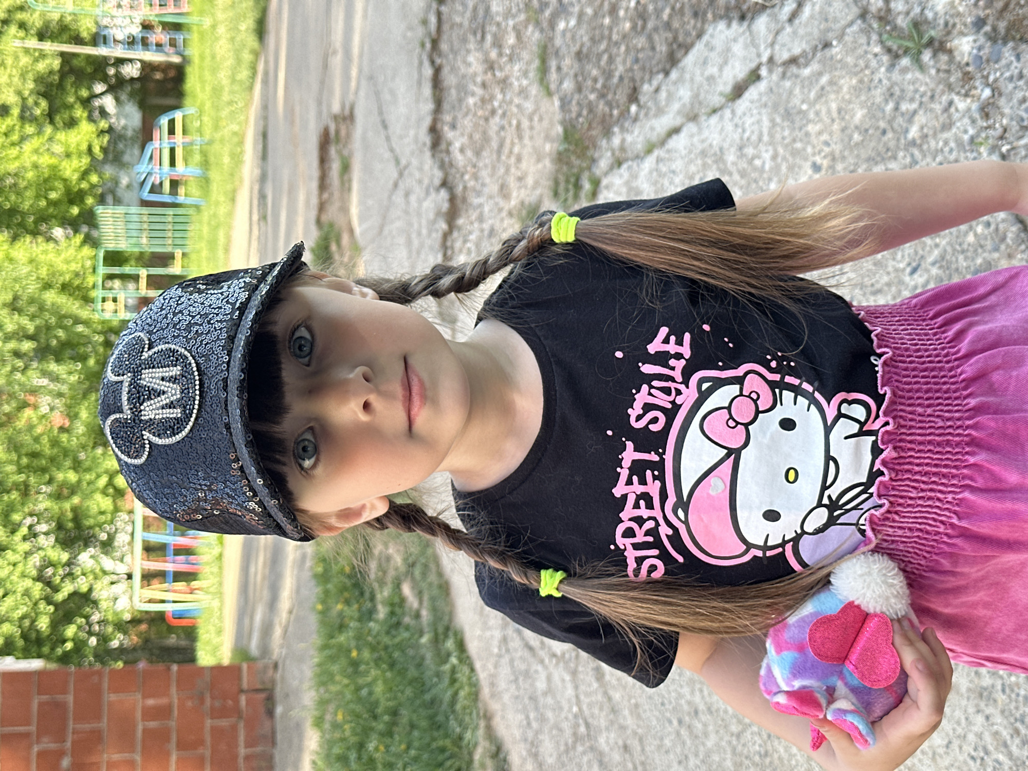 A young girl with fair skin, light brown hair styled in two braids secured with neon green bands, and light-colored eyes looks directly at the camera. She wears a dark-colored T-shirt with a graphic of Hello Kitty and the words "Street Style" printed on it, a sequined hat with a Mickey Mouse emblem, and a pink skirt. She holds a small, fluffy toy with heart-shaped decorations. The location appears to be outdoors, possibly a park or residential area, with greenery, a playground structure in the background, and a cracked concrete surface underfoot. The scene is in Chaikovsky, Russia.