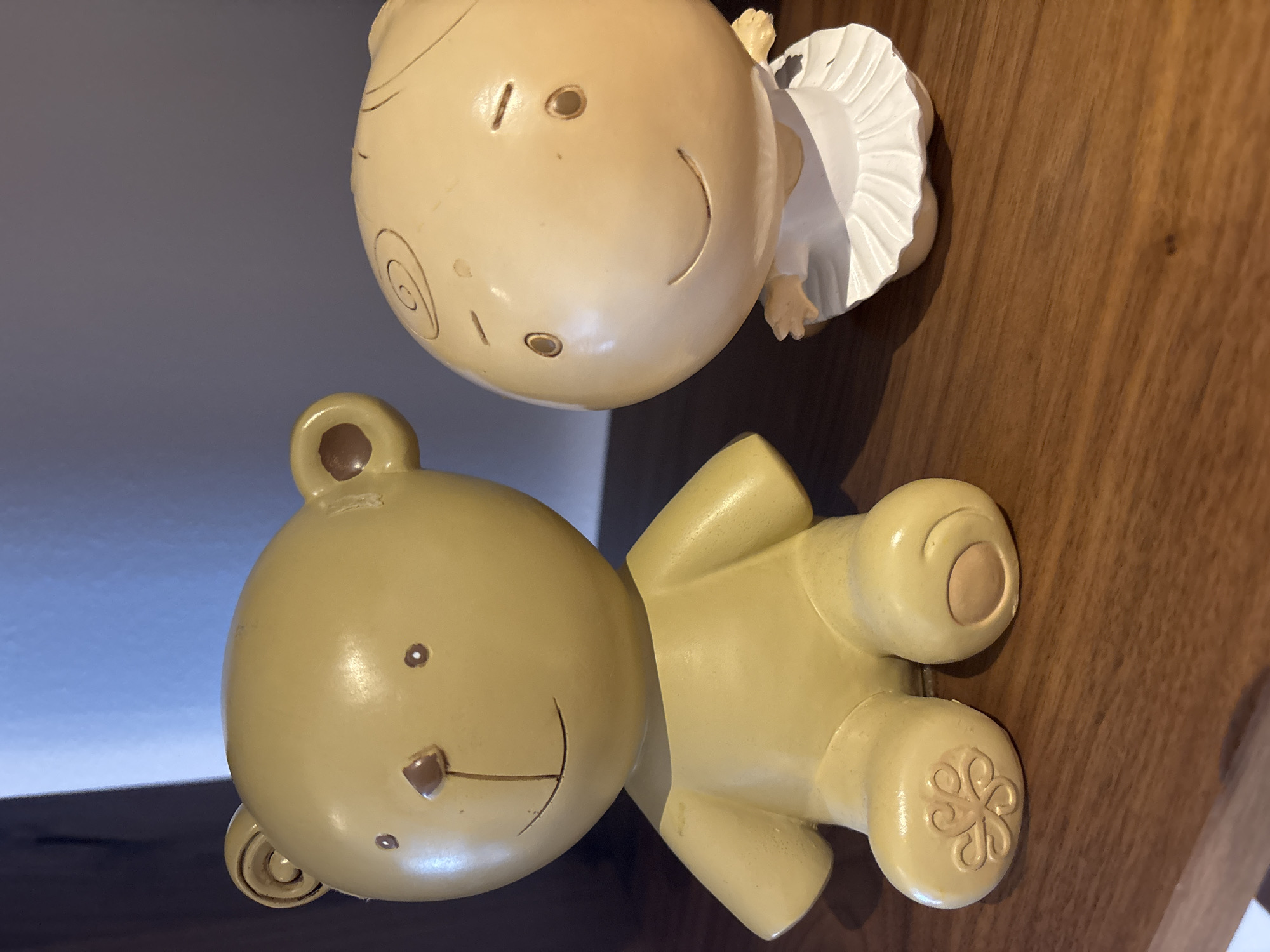 Two decorative figurines are displayed on a wooden shelf. The figurine on the left is a yellow teddy bear with small, dark eyes, a triangular nose, and a simple smile. It has round ears and a rounded body with stubby limbs. The sole of its left foot has an embossed floral or swirled pattern. The figurine on the right is a pale yellow, round-headed figure resembling a baby or cherub. It has small, round eyes, a curved smile, and is depicted wearing a white, ruffled dress or tutu. The background consists of a wall, partially illuminated by natural light. The image is a close-up, studio-style shot of these items. There is no indication of location in Dubai or the United Arab Emirates.