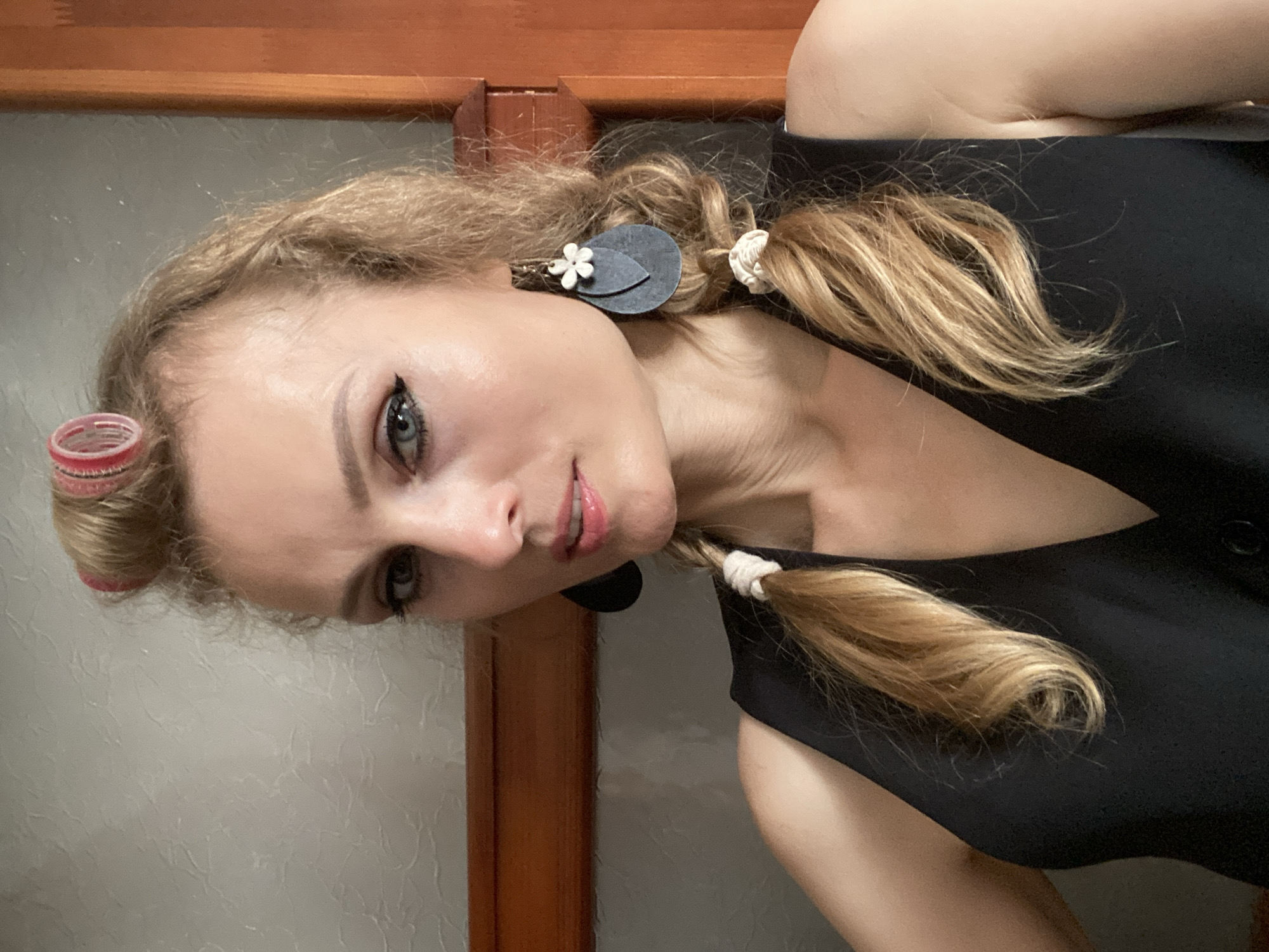 A woman with blonde hair styled in two braids and hair rollers on top of her head, wearing a black top and large, dark, teardrop-shaped earrings with a white flower embellishment. She is indoors, against a textured, light-colored wall and a wooden frame.