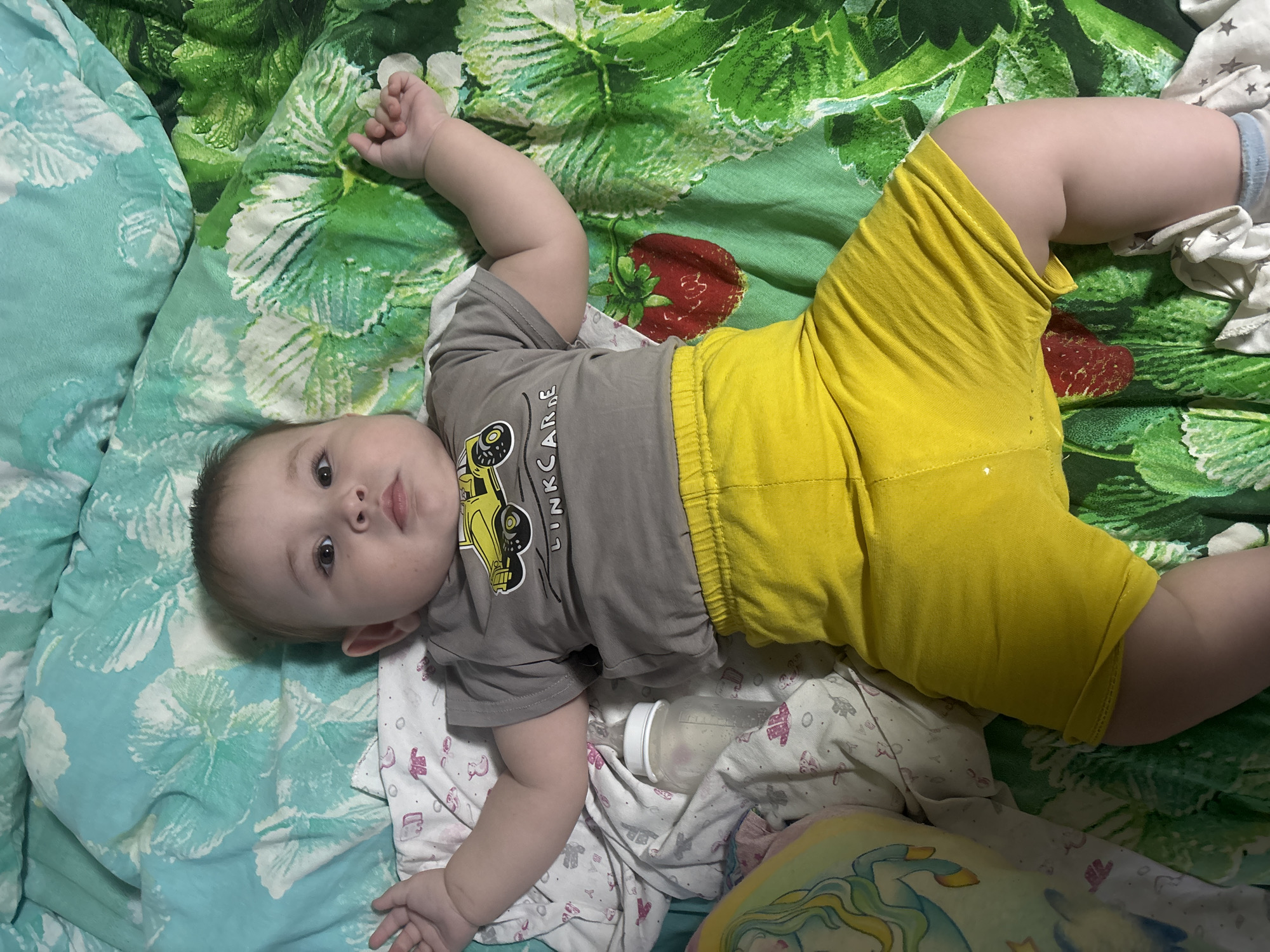 An infant, appearing to be several months old, is lying supine on a bed, positioned centrally in the frame. The infant is dressed in a short-sleeved grey t-shirt featuring a yellow graphic of a construction vehicle and partially visible text "LINK CAR," paired with bright yellow elasticated shorts. A blue and white striped sock is visible on the infant's left foot (viewer's bottom right).
The infant is positioned on multiple layers of bedding. Immediately beneath is a white sheet with repeating pink motifs, including baby clothing, ducks, and the word "BABY." This layer rests on a duvet or blanket with two distinct patterns: a light blue floral/leaf pattern on the upper left, and a green background with dark green leaf patterns and prominent red strawberry illustrations covering the rest of the visible surface. To the infant's left side, a partially visible clear baby bottle with a white cap containing a small volume of liquid is present, resting on the white patterned sheet. Adjacent to the infant's head on the left, a multi-colored object, possibly a pillow or soft toy, displays a light blue and yellow unicorn/Pegasus design.
The infant's gaze is directed forward, towards the observer/camera, with an alert and neutral facial expression. Both arms are slightly flexed and abducted, with hands open. Legs are flexed at the hips and knees, with feet splayed.
The image itself contains no visual information or metadata that specifies a geographic location such as the city of Chaikovsky, Russia.