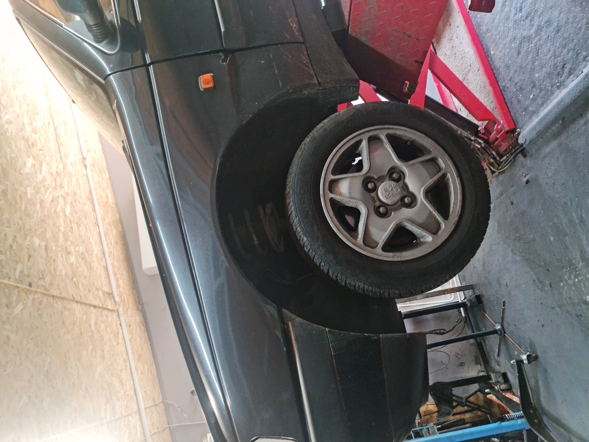 This image captures a dark-colored car, likely a hatchback or small SUV, elevated on a red hydraulic lift within what appears to be an automotive workshop or garage. The car is positioned at an angle, with its rear section and wheel prominently displayed.
The vehicle's paintwork is a dark gray or dark green metallic, and an amber side marker light is visible on the rear quarter panel. Distinct fender flares are noticeable above the wheel. The rear wheel features a silver, five-spoke alloy rim, with a central hubcap bearing a logo that resembles a lion emblem, possibly indicating a Peugeot vehicle. The tire's sidewall clearly displays the brand name "SYRON."
The red structure of the car lift is visible, with its metal frame and some hydraulic components extending towards the floor and supporting the car. Beneath the elevated car, the concrete floor is visible, showing areas of grime and wear, along with various tools, cables, and equipment, including parts of a blue jack or stand.
To the left of the car, a wall covered with light-colored wooden panels, resembling OSB or chipboard, is present, with white electrical conduit running across it. The overall setting strongly suggests an indoor automotive repair or maintenance activity taking place. No people are visible in the scene. The location context provided is Buzovgrad, Bulgaria.