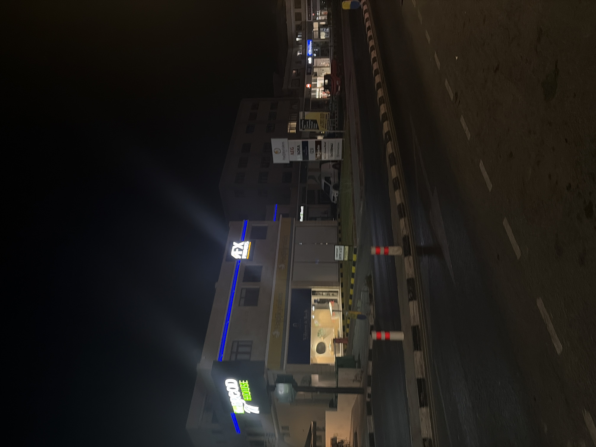 This is a night-time, eye-level, medium shot of a street scene in Agios Athanasios Municipality, Cyprus. The primary focus is on a building with illuminated signage, suggesting a commercial or office space. The building features a prominent green neon sign with "MERIGOD HOUSE" and the number "7". Adjacent to it, another sign reads "AFX FINANCIAL SERVICES" in white lettering. Below this, a yellow awning stretches across the storefronts. One storefront is visible with the name "Villages & Bach" displayed, and the interior is lit, showing displays or furniture.
Further down the street, a tall signpost displays multiple logos and brand names, including "HELLAS TRADING GROUP", "AEG", and "BORA". Several other storefronts are visible with their own illuminated signs. A white van and a red truck are parked in front of some of these establishments.
The street itself is paved, and there are traffic bollards with red and white reflective markings lining the pavement. The road has visible white lane markings. The overall environment is dark, illuminated primarily by the building lights and streetlights, creating a nighttime urban atmosphere. No people are clearly visible in the scene.
