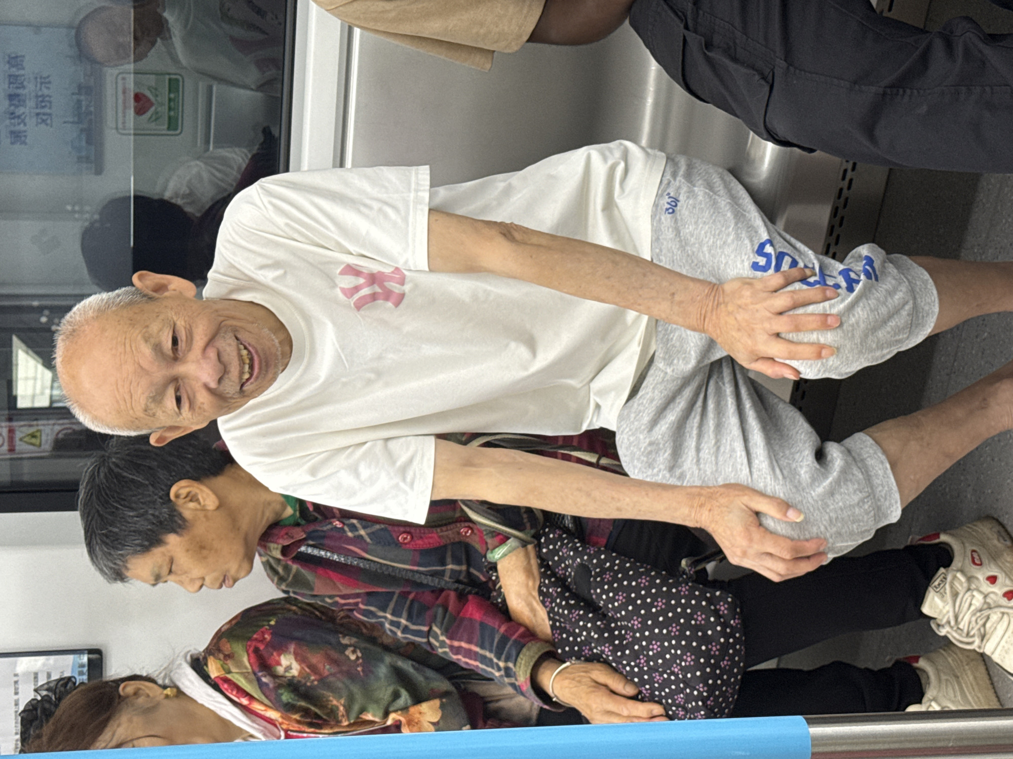The image depicts a group of people, predominantly elderly, seated on a subway train. The central figure is an elderly Asian man with a bald head and a wide smile, wearing a white t-shirt with a pink logo and grey sweatpants. To his left, an elderly Asian woman with short dark hair wears a plaid shirt and has a patterned bag on her lap. Another woman, partially visible on the far left, has dark hair and wears a colorful jacket. To the right of the central figure, another person is visible wearing a tan t-shirt and black pants. The environment is the interior of a subway car, with metal seating and reflections in the window. The text in the background appears to be in Chinese. The scene is captured in what is identified as Tiaodeng, China.