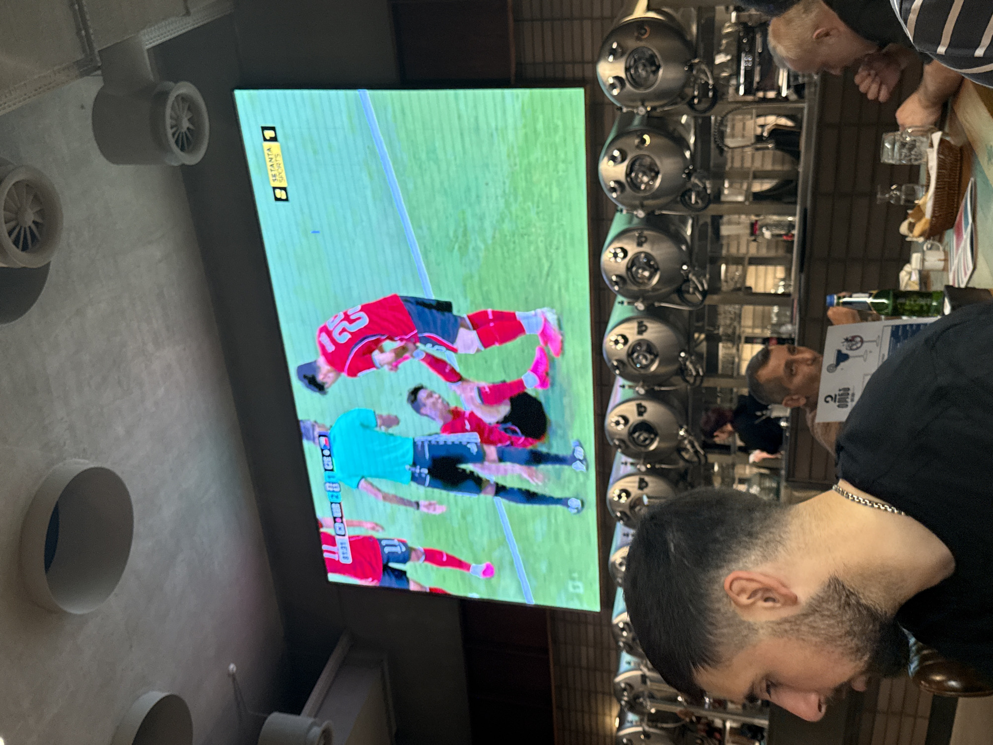 The image depicts a dimly lit interior of what appears to be a bar or pub, with a large screen displaying a soccer match. Several patrons are visible, some seated at tables in the foreground and others in the background. The screen shows two teams playing, with one player on the ground and another standing over him. A score graphic is visible on the screen, indicating "SETANTA SPORTS 1". In the foreground, a man with dark hair and a beard is looking to his left. Behind him, a bar area with several chrome tanks or kegs is visible, along with a bartender or staff member in the background. The overall atmosphere suggests a casual social gathering centered around watching a sporting event.